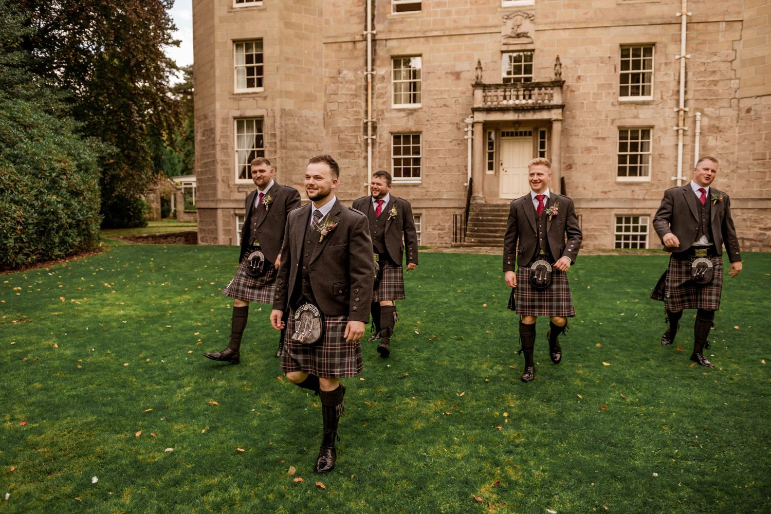 Groomsmen in traditional Scottish kilts pose on grass in front of historic stone building.