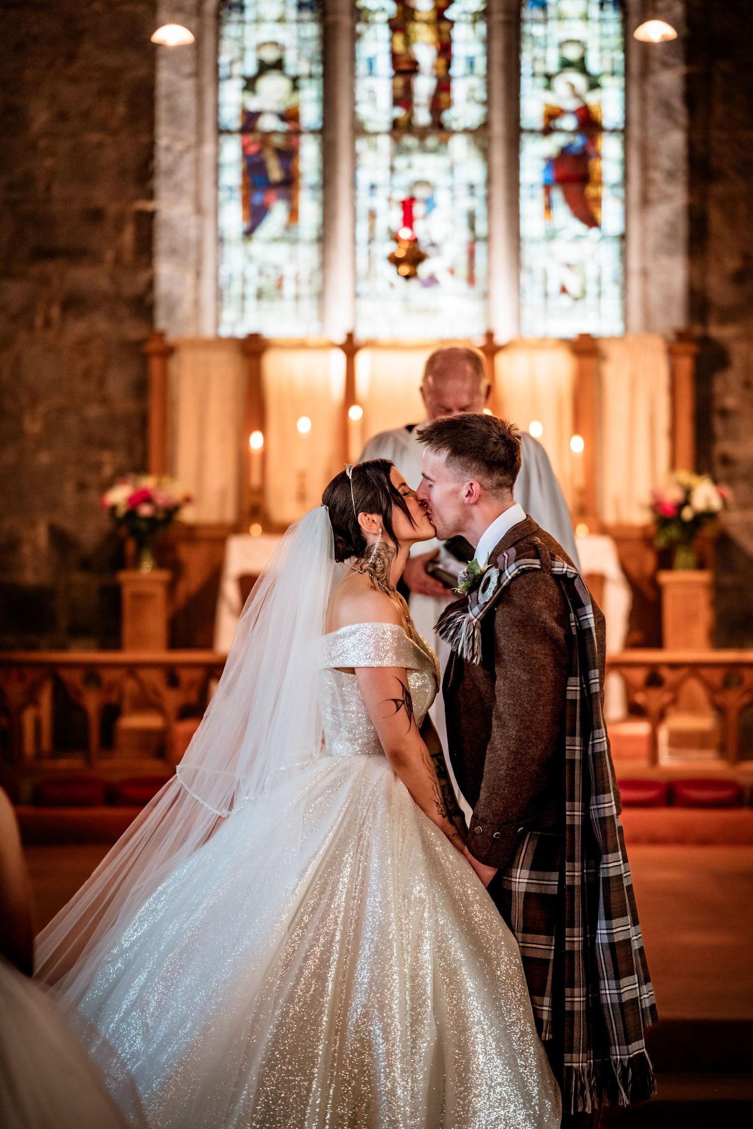 A romantic wedding kiss in a church with stained glass windows and glowing candles in the background.