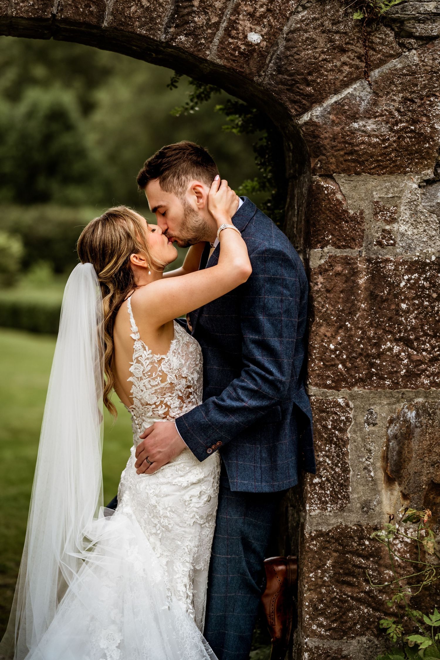 Bride and groom share a romantic kiss under a stone arch, bride wearing lace gown with veil, groom in navy suit.
