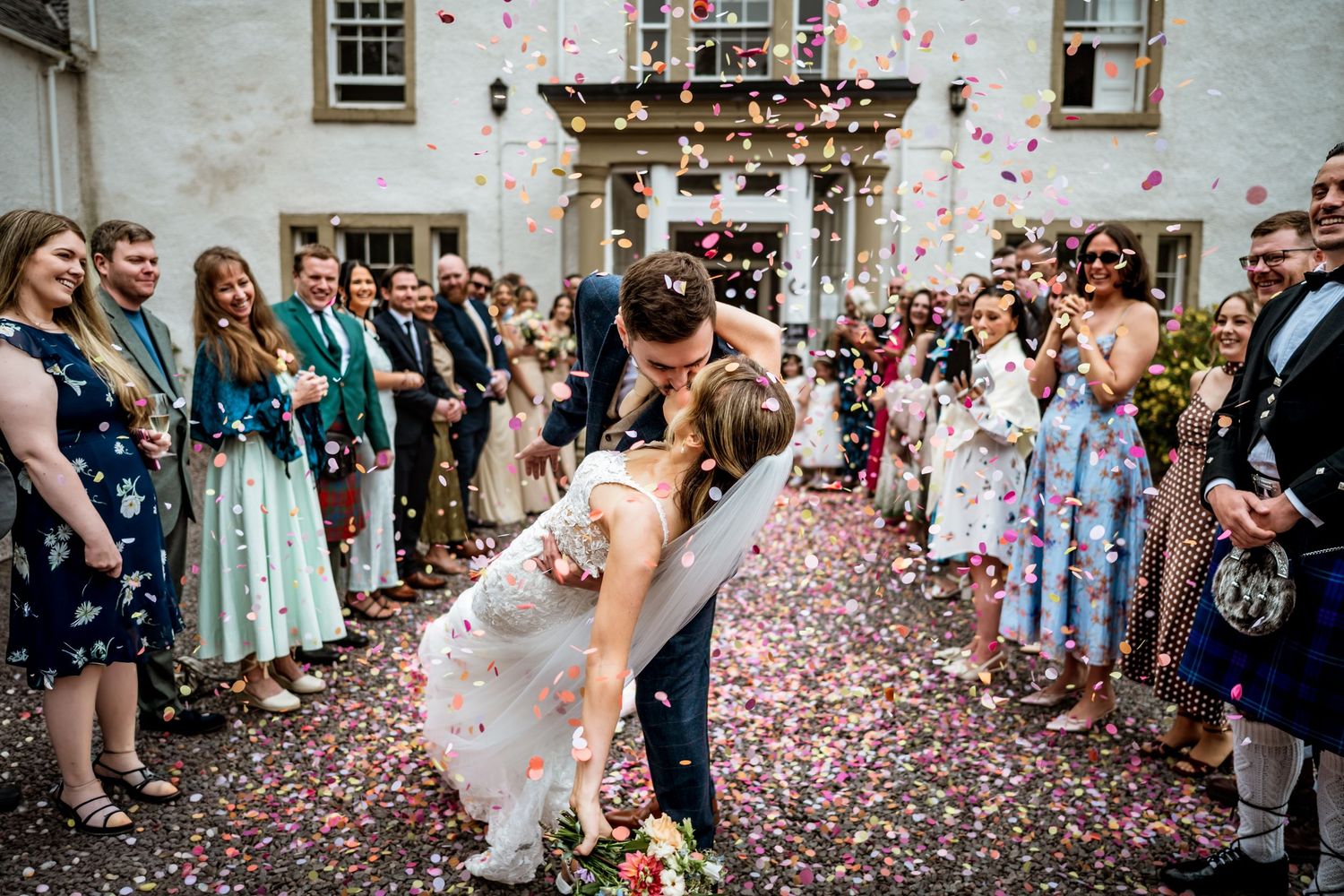 Bride and groom share romantic dip kiss surrounded by colorful confetti and cheering wedding guests outside elegant venue.
