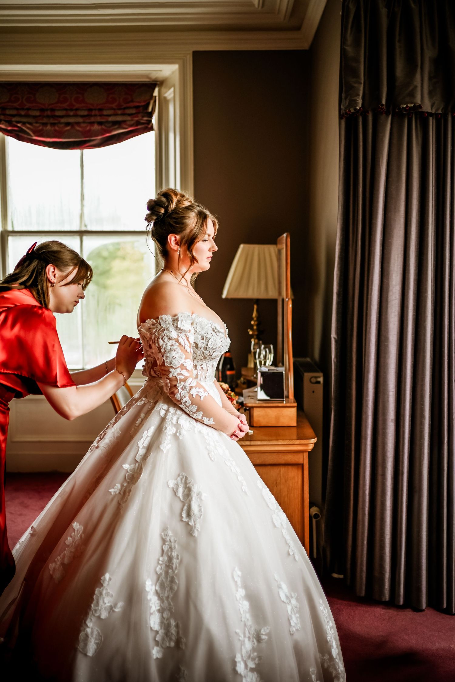 Side view of stunning white lace wedding gown being adjusted in elegant room.