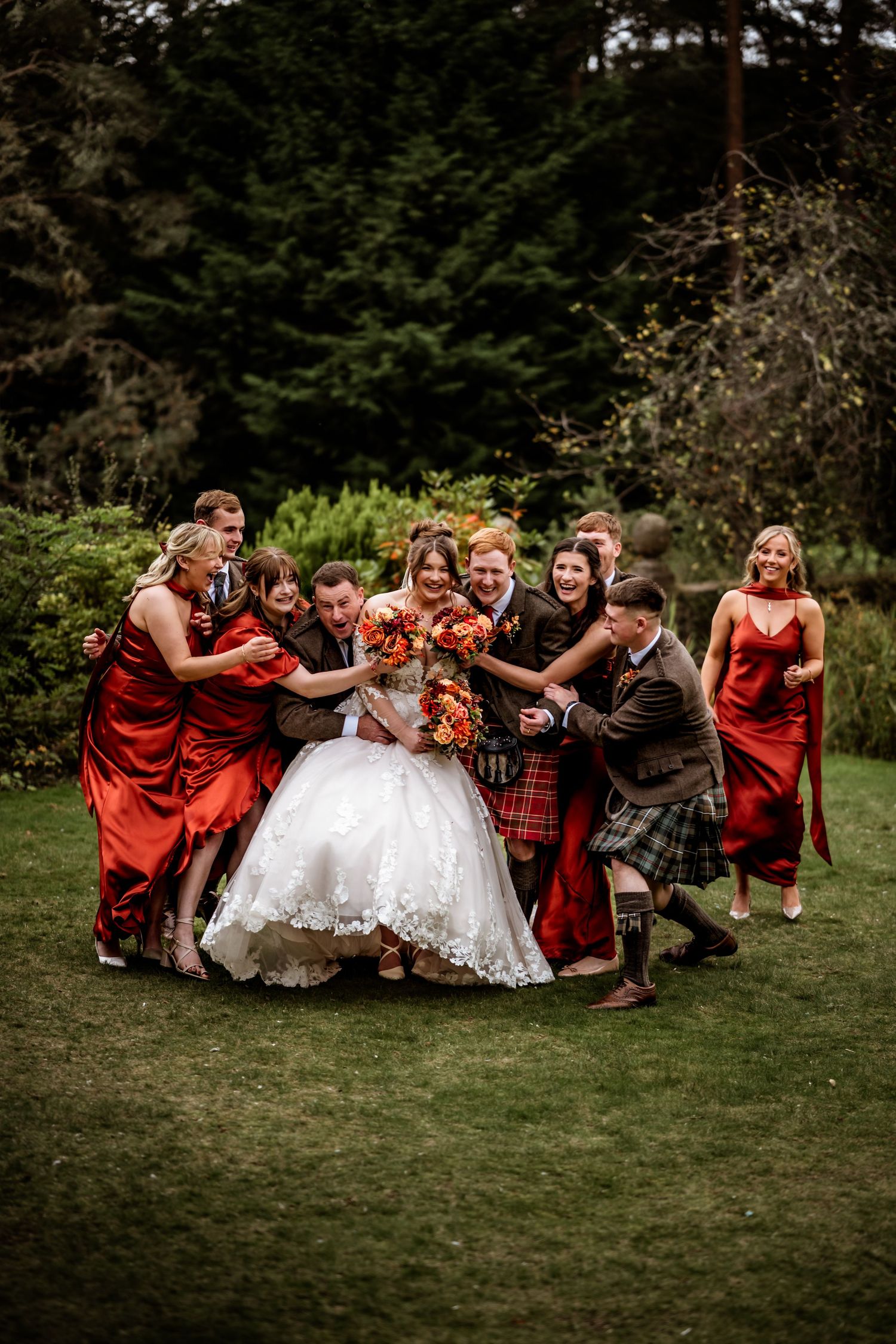 Wedding party in red dresses and kilts poses playfully on grass for a fun group photo.