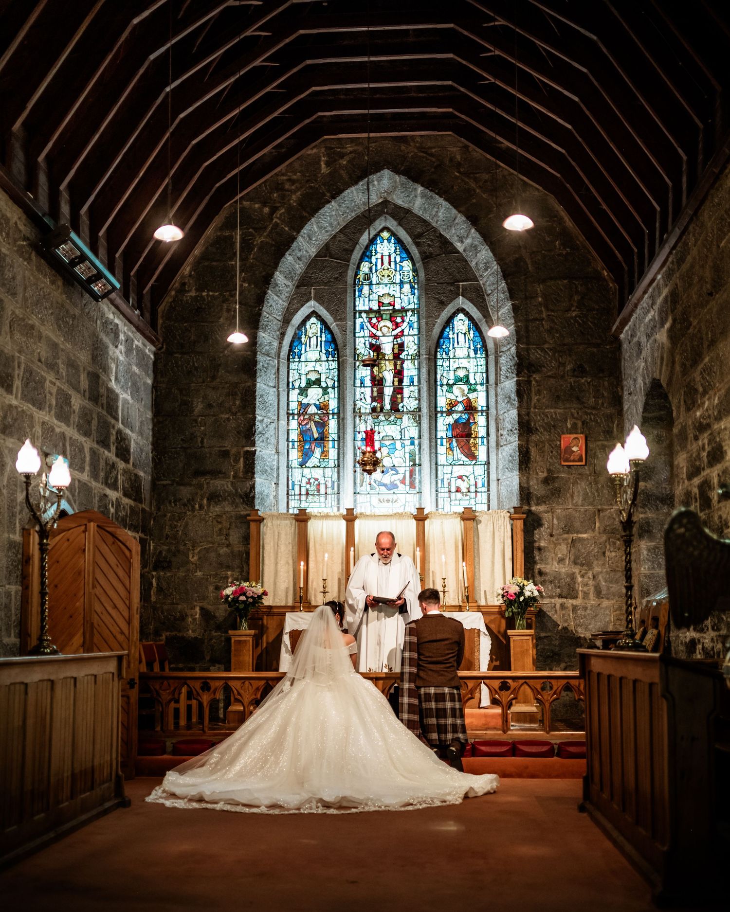 A bride in flowing white gown stands before ornate stained glass windows in historic church.