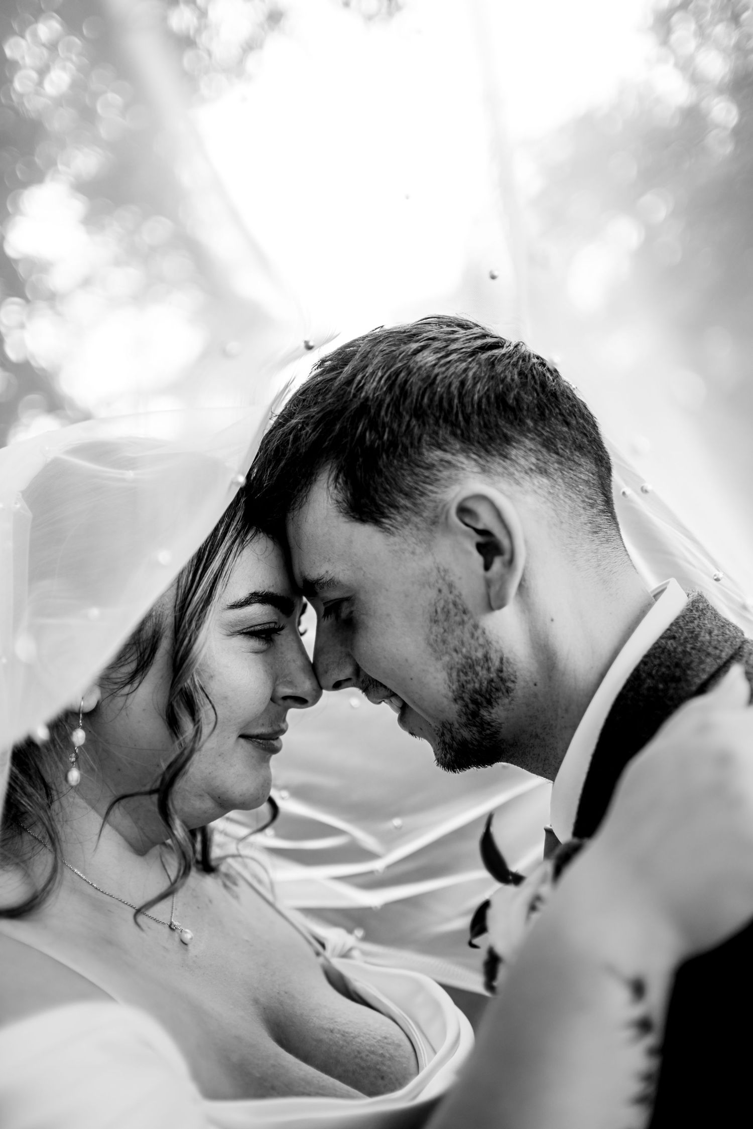 Black and white photo of bride and groom touching foreheads tenderly under a veil on their wedding day.