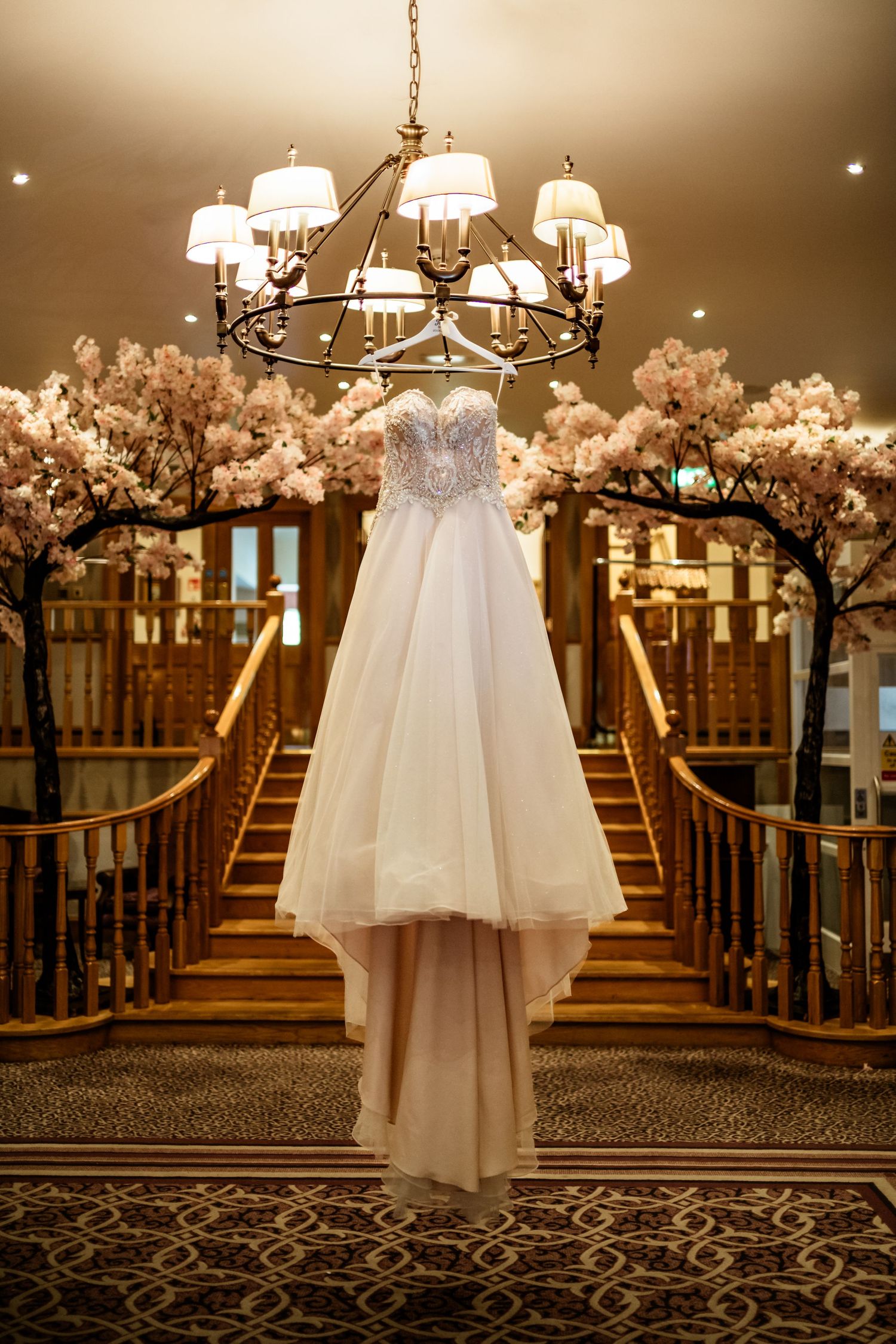 Elegant beaded wedding dress hanging from a chandelier above grand staircase with cherry blossom trees in luxurious venue.