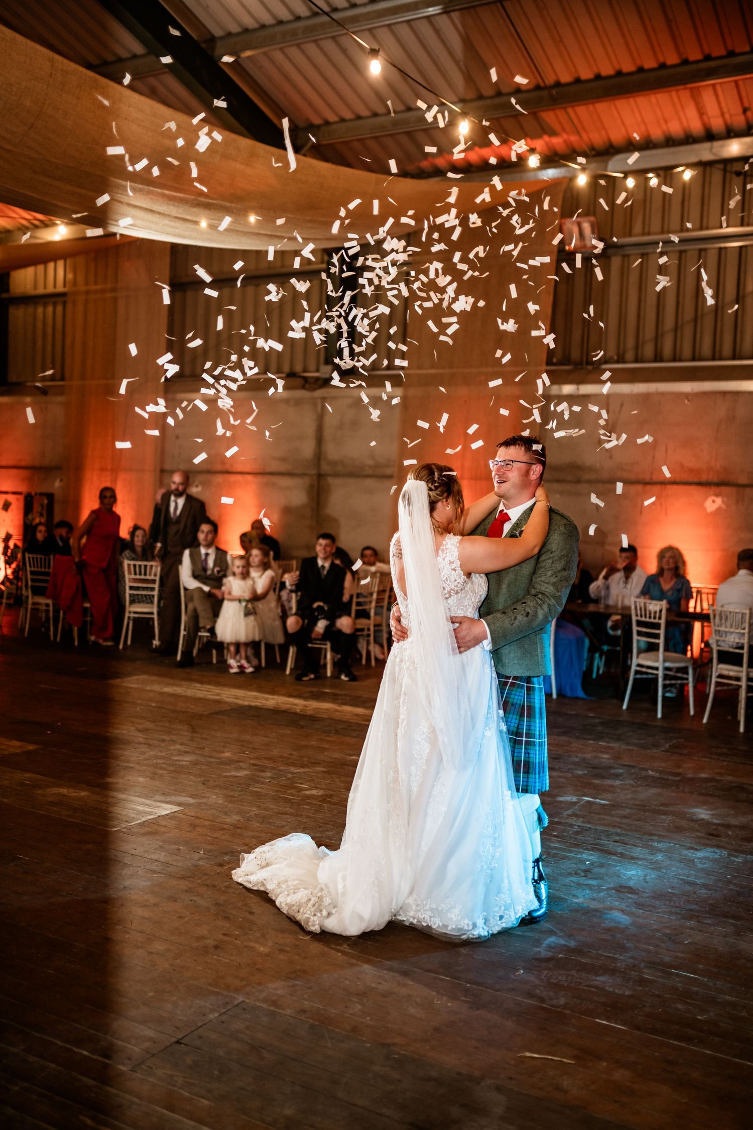 A wedding couple's first dance with confetti falling in a rustic venue with warm lighting. Scottish Highlands Farm Wedding photography