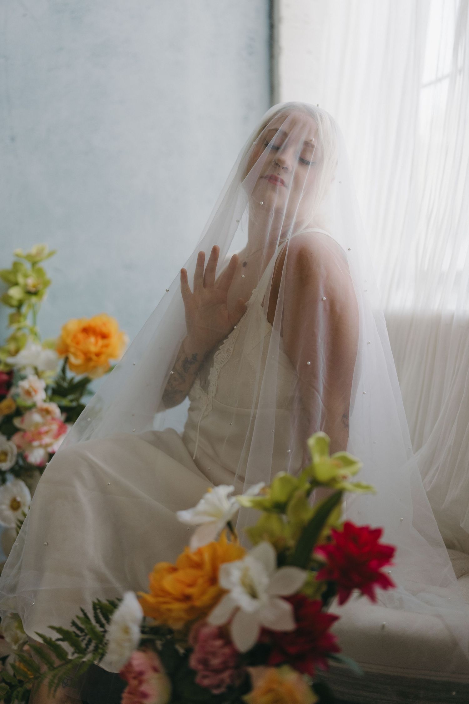A bride in a white gown and pearl-dotted veil poses gracefully among colorful floral arrangements in a studio setting in Minneapolis, MN.