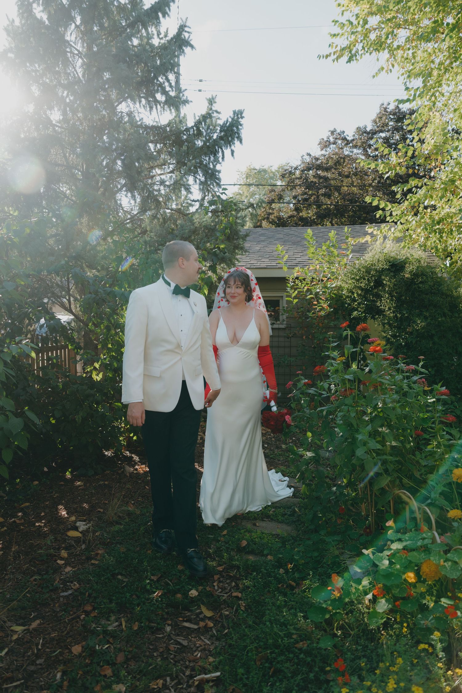 Bride and groom walking through lush garden in sunlight, she wears red gloves near Minneapolis.