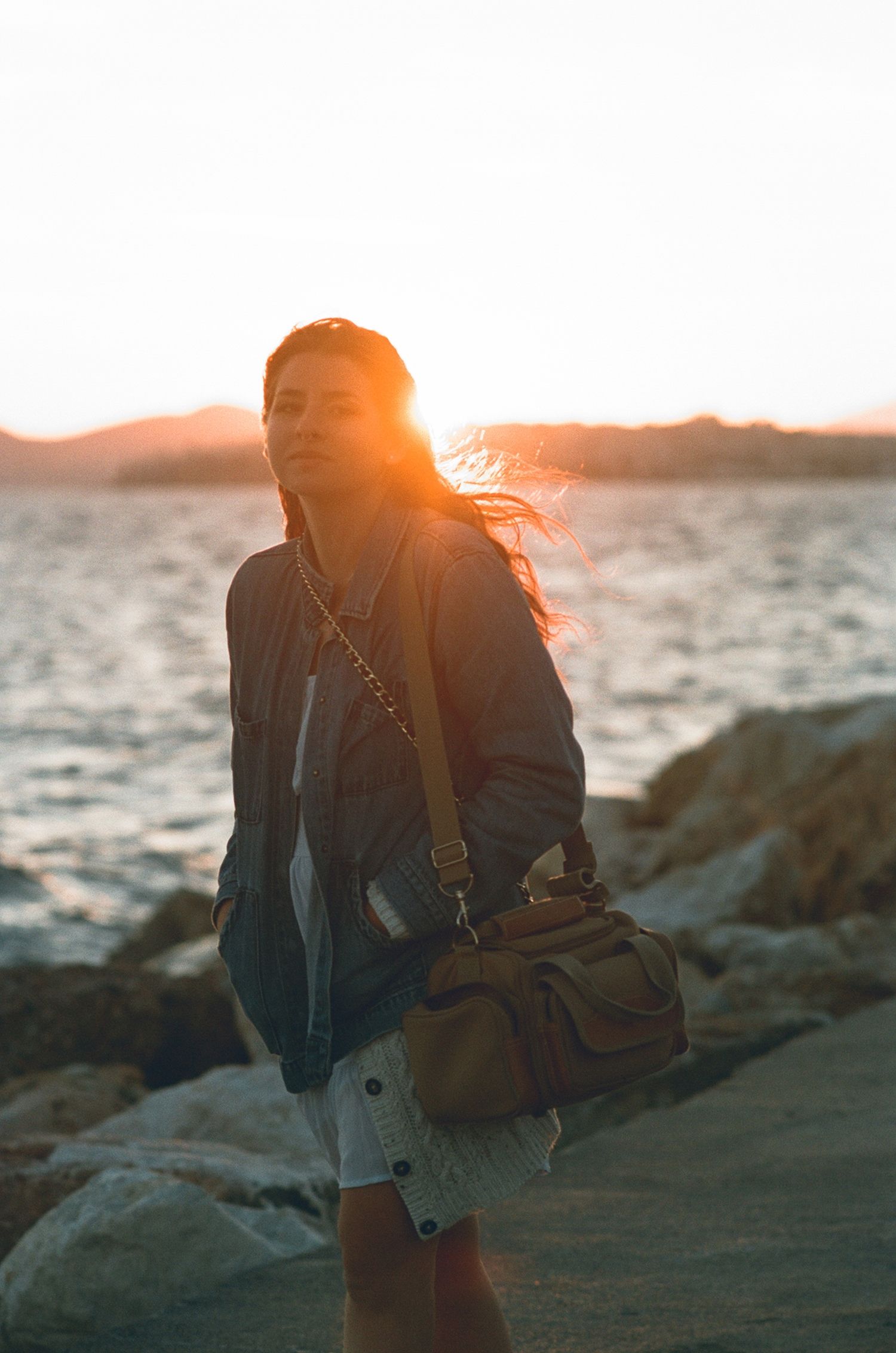 Woman with shoulder bag stands on rocky shoreline with hair blowing in golden sunset light in Greece shot on 35mm film.