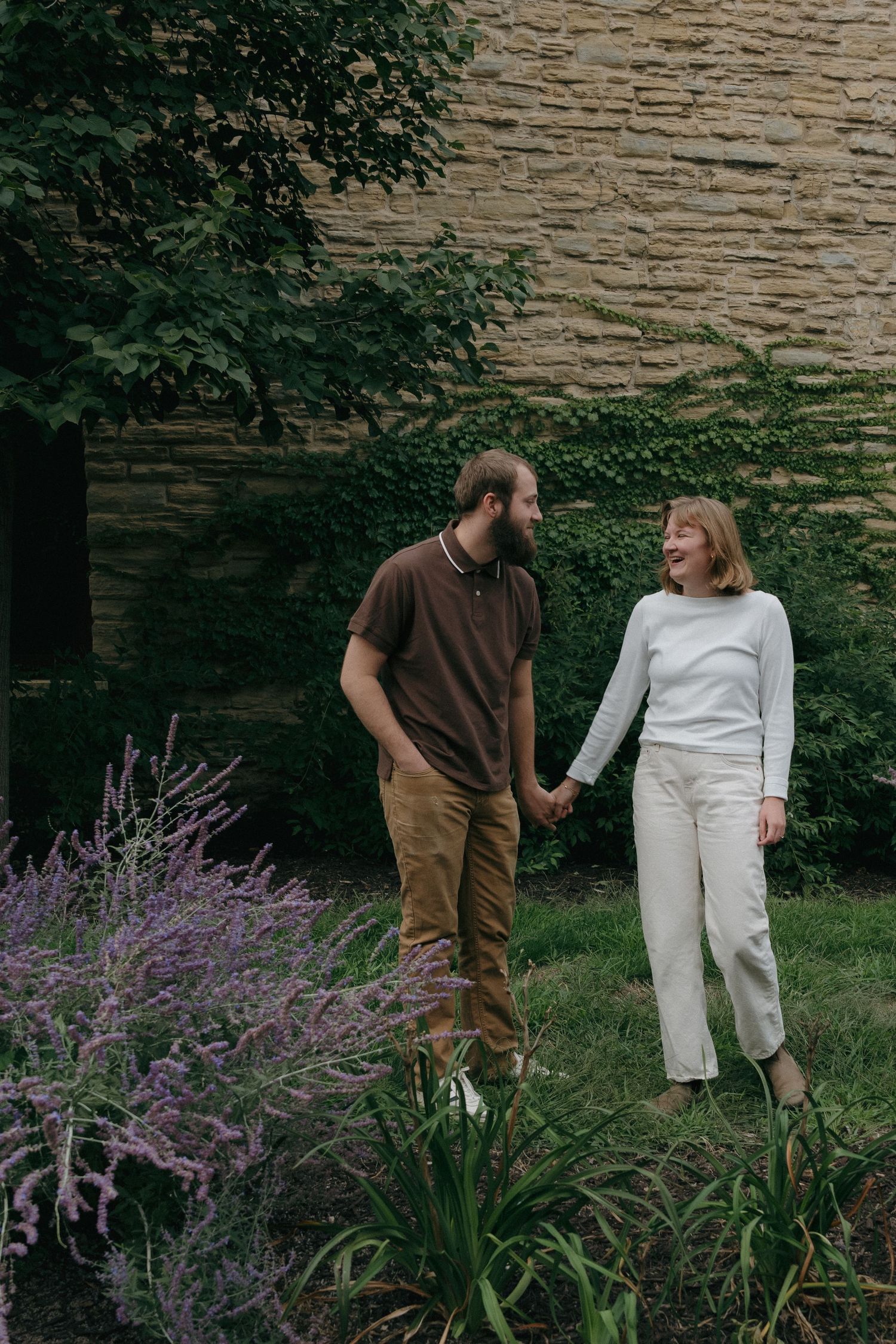 Happy couple holding hands and laughing together in a lush garden with purple flowers and stone wall near Saint Anthony Main in Minneapolis.