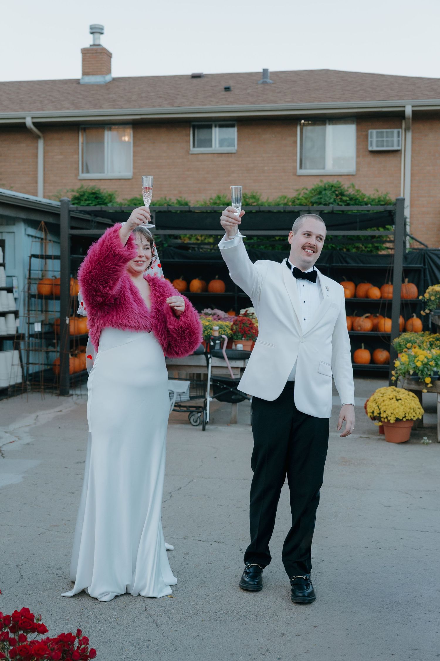Bride in white gown with pink fur shrug and groom in white tuxedo toasting champagne outdoors.