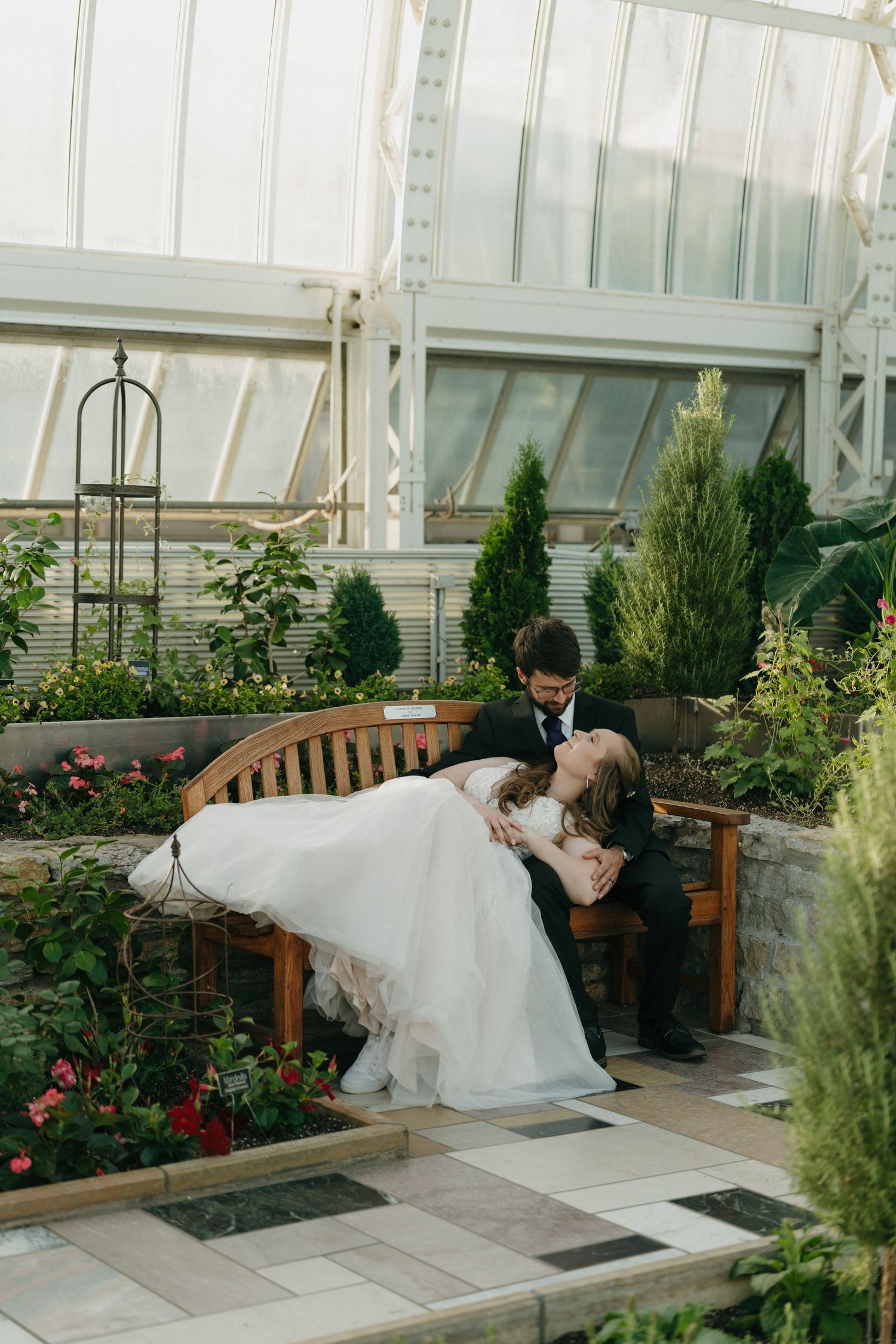 Bride and groom share a romantic dip kiss on a wooden bench inside a lush greenhouse wedding venue.