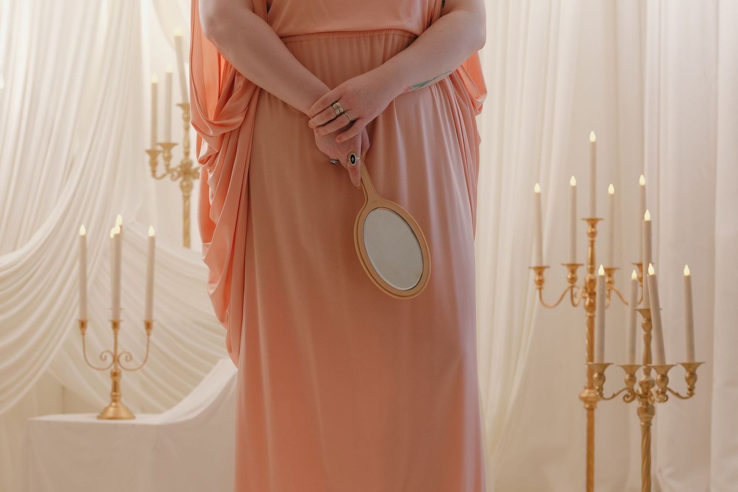 Woman in peach flowing gown holding a gold oval hand mirror beside golden candelabras in a studio in St. Paul, Minnesota.