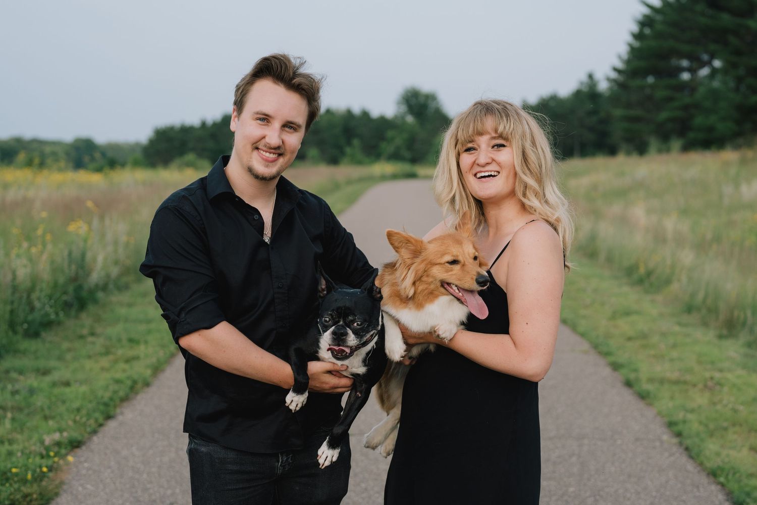 Smiling couple in matching black outfits holding two dogs on a scenic path at Schaar's Bluff Regional Park in Hastings, Minnesota.