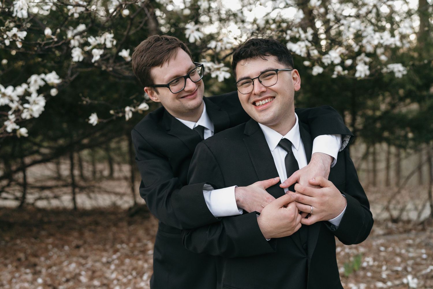 Two grooms in black suits share a joyful embrace outdoors near blooming white flowers.