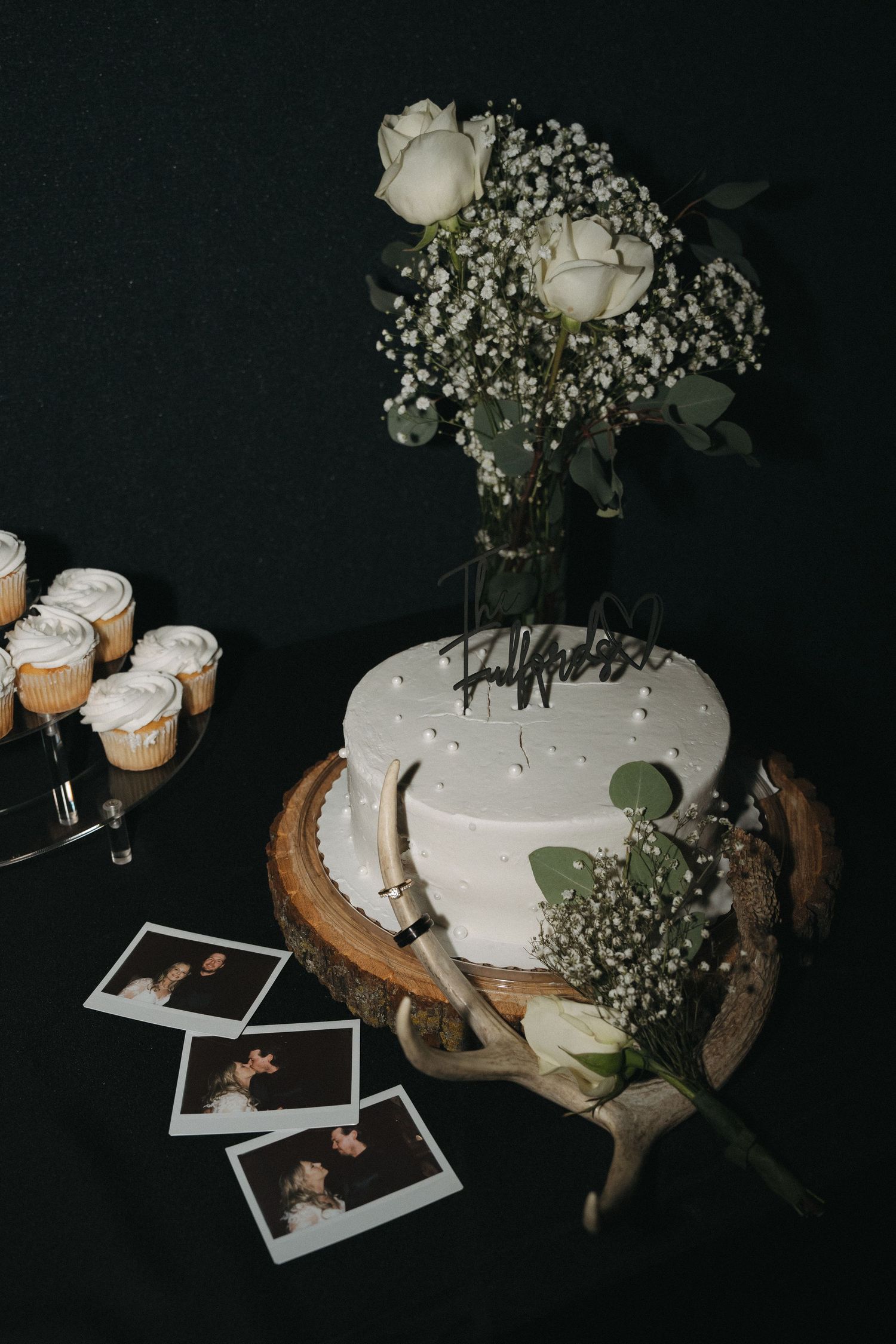 Elegant white wedding cake on wood slice with roses, baby's breath, antlers, and Polaroid photos at a wedding reception.