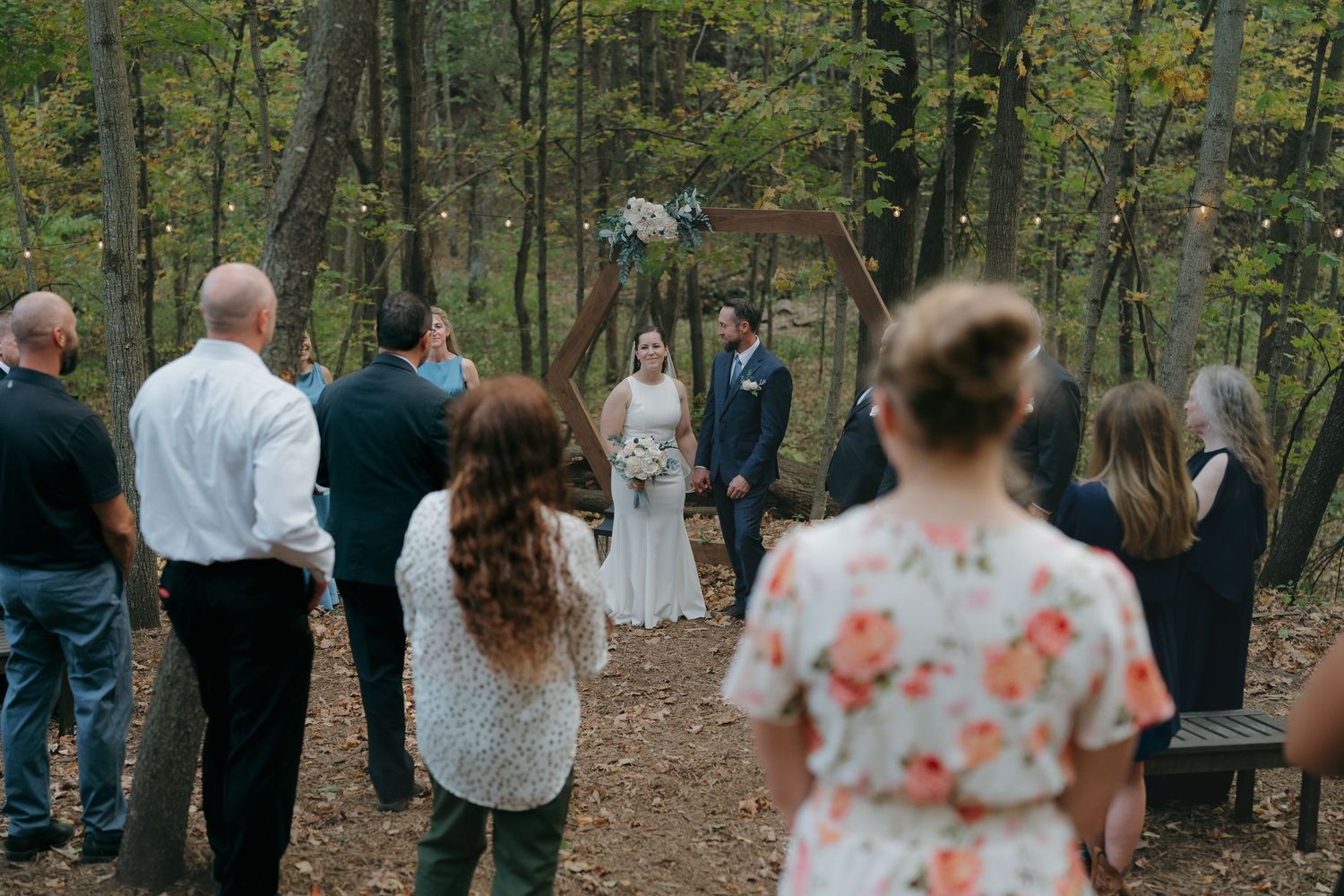Outdoor woodland wedding ceremony with guests watching bride and groom at a rustic wooden arch altar in Southern Minnesota.