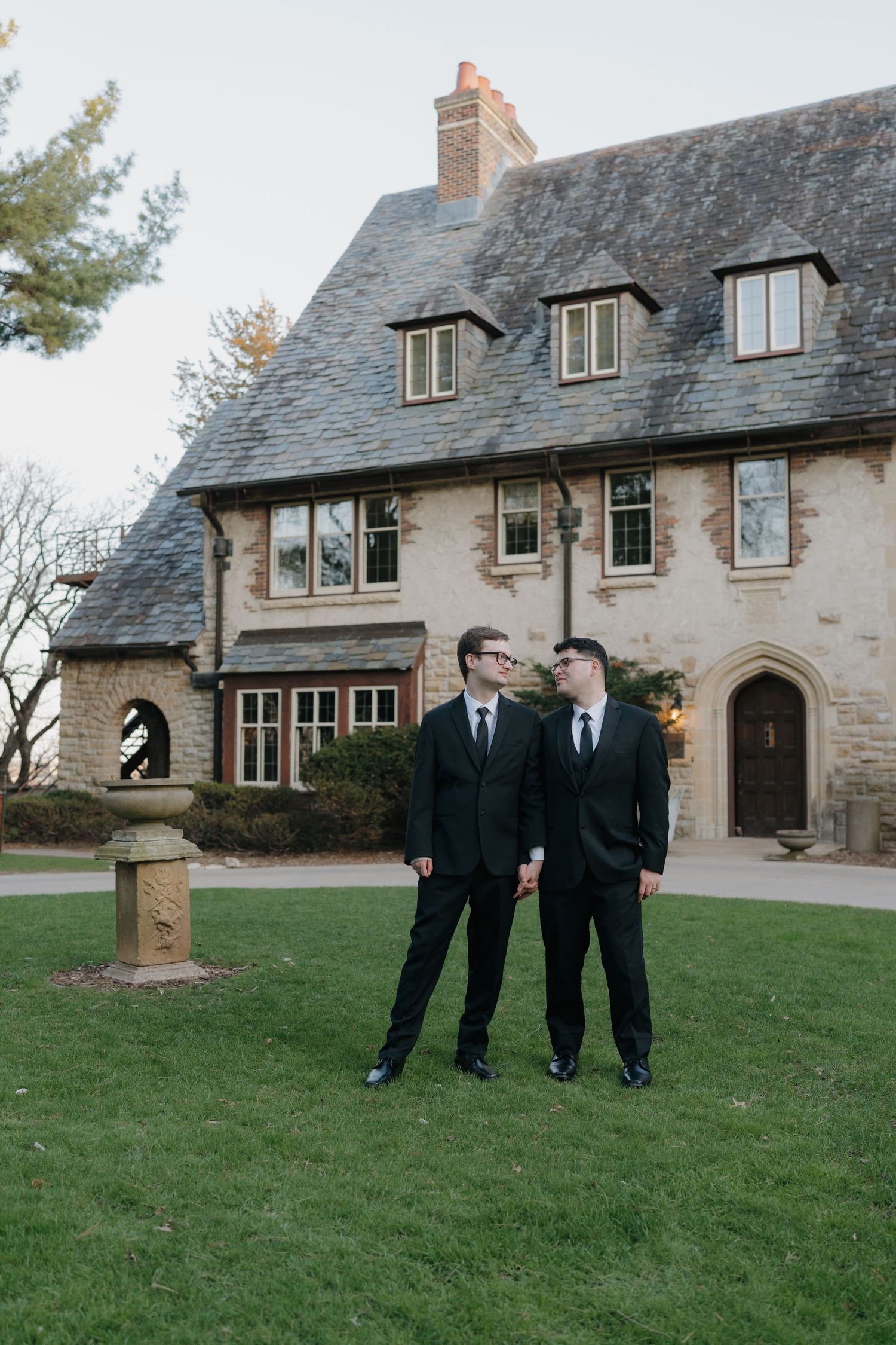 Two grooms in black suits stand together on a lawn before an elegant stone manor house at dusk.