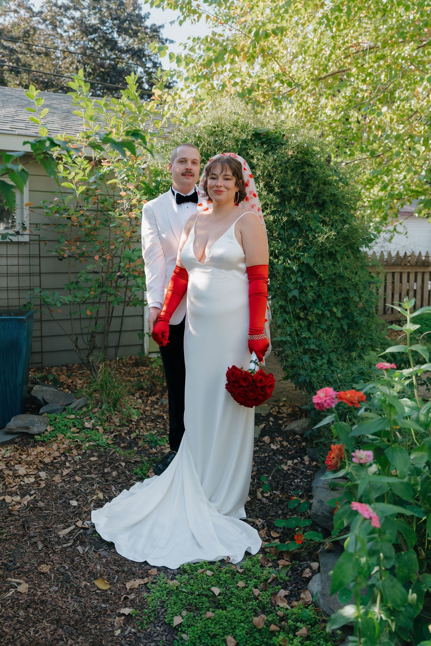 Bride in white gown with red gloves holds red bouquet beside groom in white tuxedo in garden in Minneapolis, Minnesota.