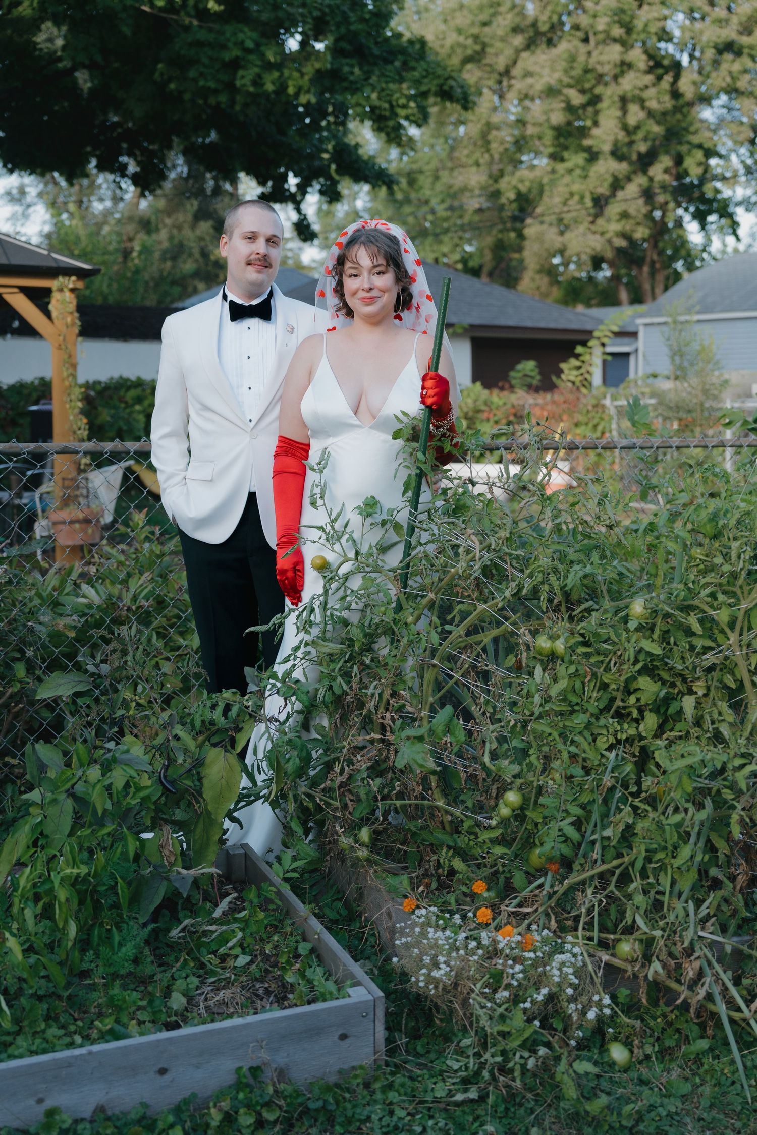 Wedding couple posing in a lush garden, bride wearing red gloves holding a fence post in Longfellow, Minneapolis, Minnesota.
