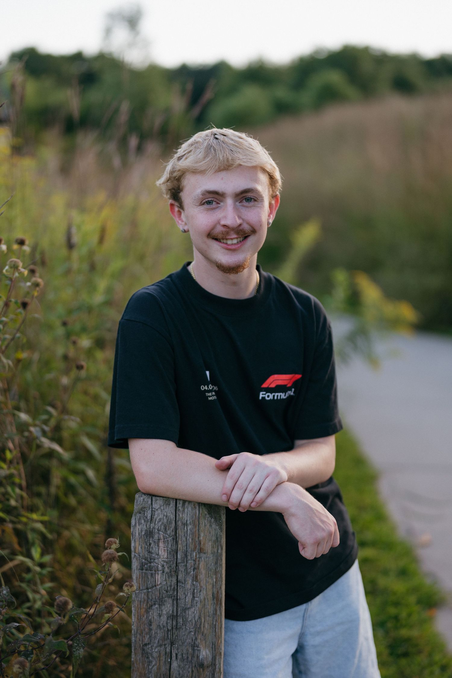 Smiling young blond man in Formula 1 black t-shirt leaning on wooden post along a nature trail in Eagan, Minnesota at Lebanon Hills.