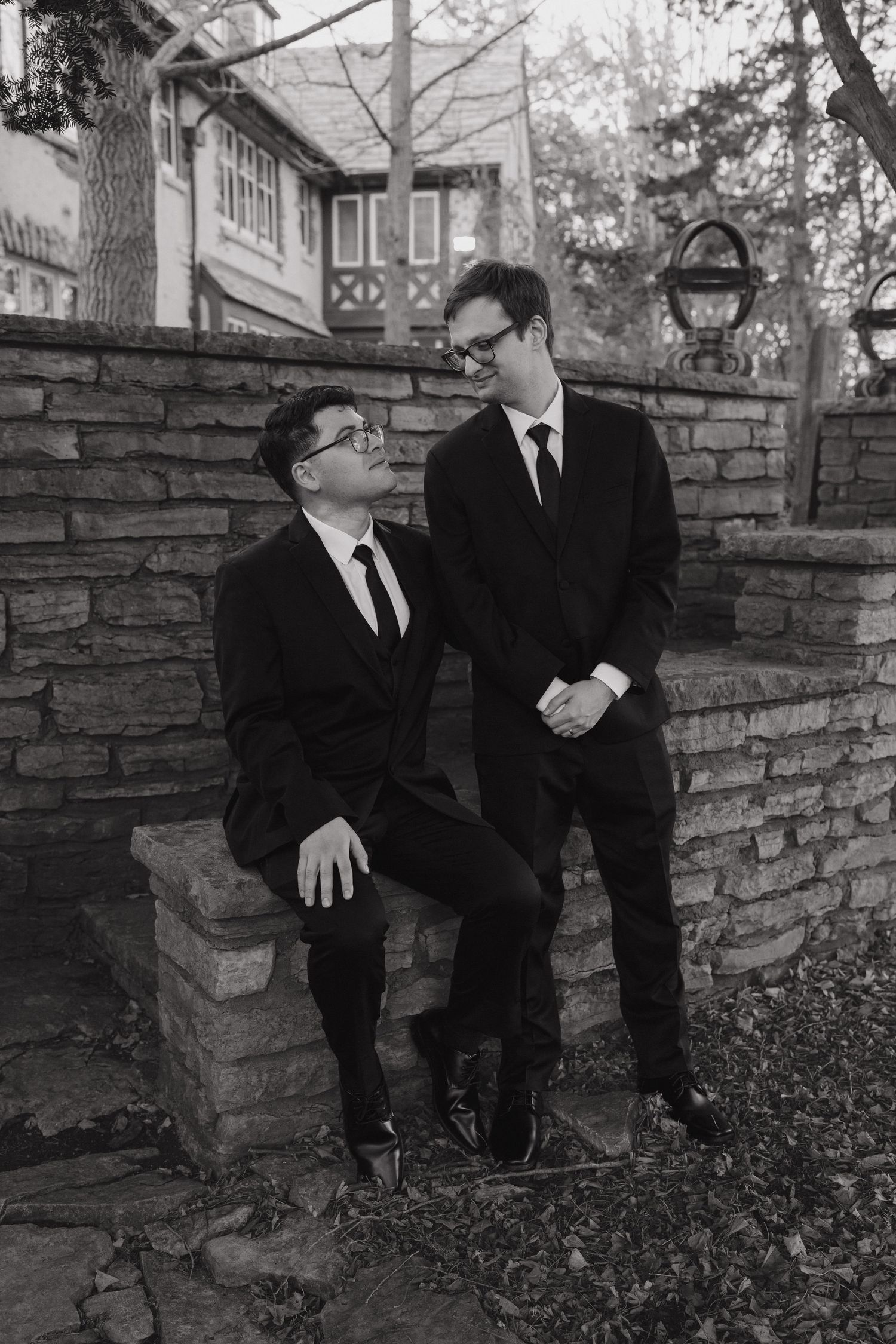 Black and white portrait of two grooms in black suits smiling together on a stone wall outdoors near Rochester, Minnesota.