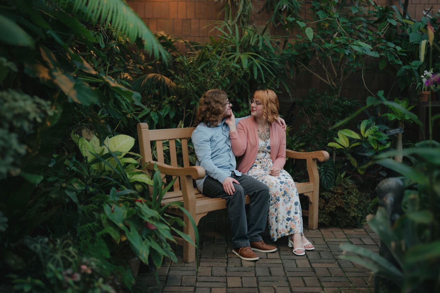 Couple sitting together on a wooden bench surrounded by lush tropical greenery at the Minnesota Landscape Arboretum greenhouse.