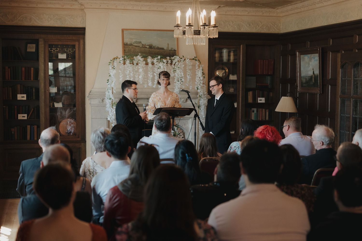Elegant indoor wedding ceremony in a library with couple exchanging vows before seated guests at Plummer House in Rochester, Minnesota.