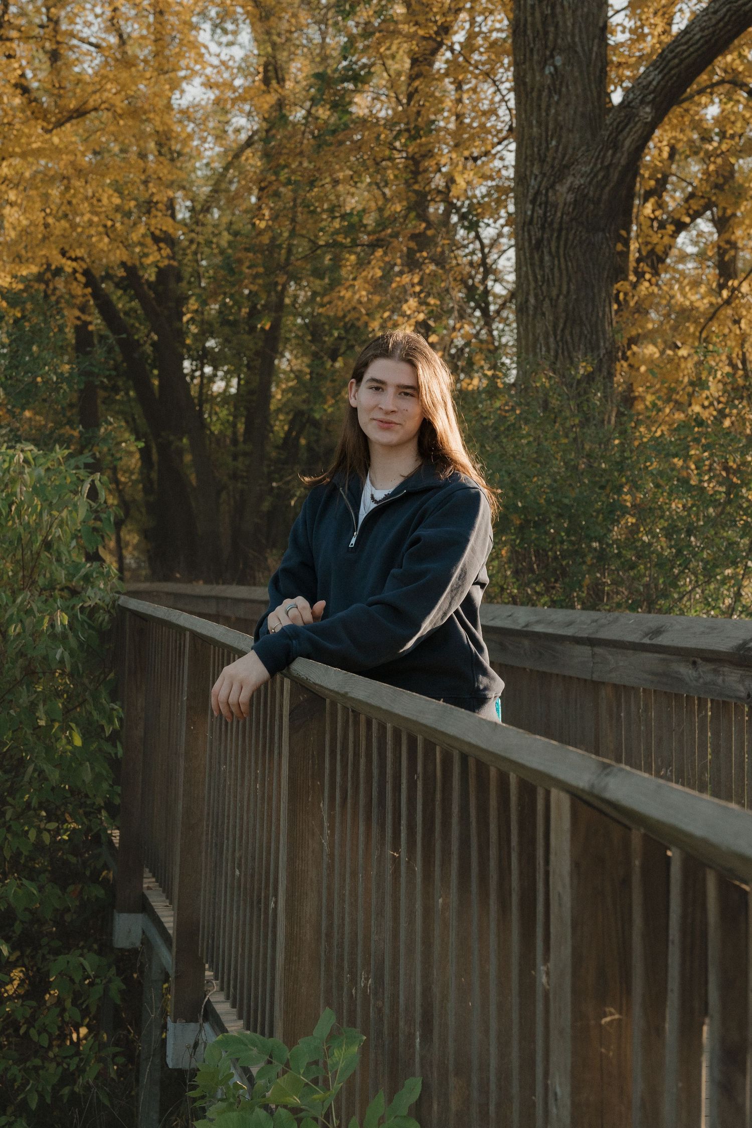 Tee in dark zip-up jacket leaning on wooden bridge railing with golden autumn trees behind them for a golden hour senior session in MN.