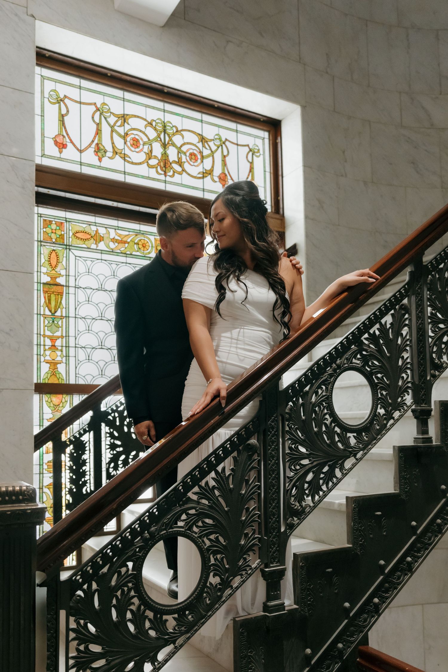 Couple shares romantic moment on ornate iron staircase beneath colorful stained glass window at Minneapolis City Hall.