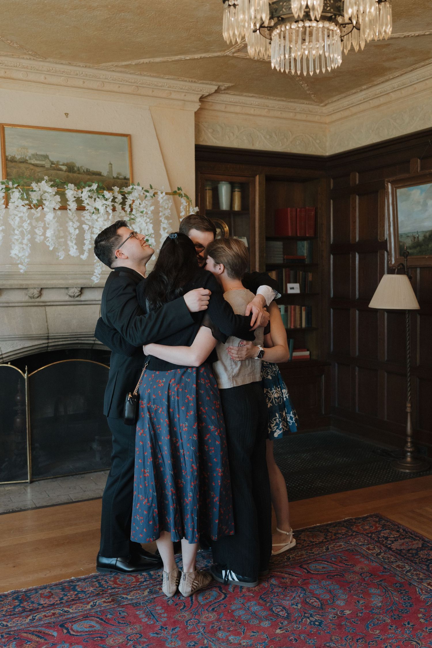 Four people sharing a warm group hug in an elegant wood-paneled room at the Plummer House with a crystal chandelier in Rochester, Minnesota.
