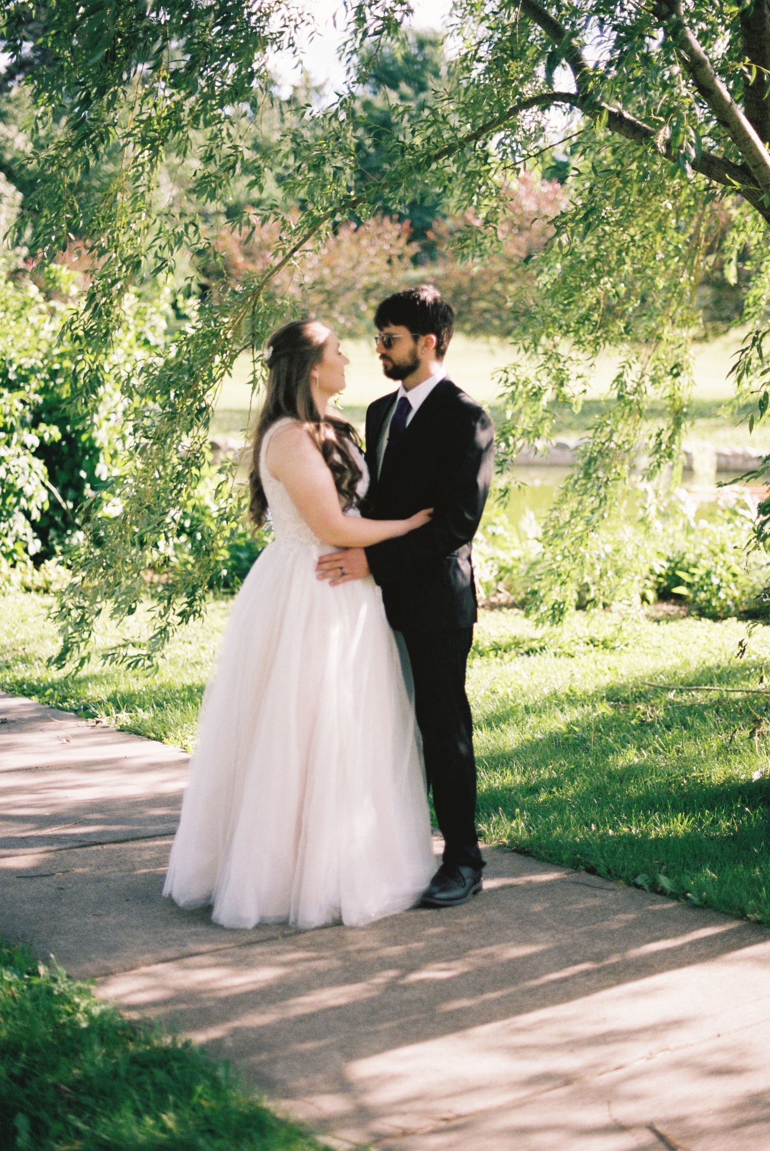 A bride in a white tulle gown and groom in a black suit share a romantic moment on a garden path surrounded by lush greenery.