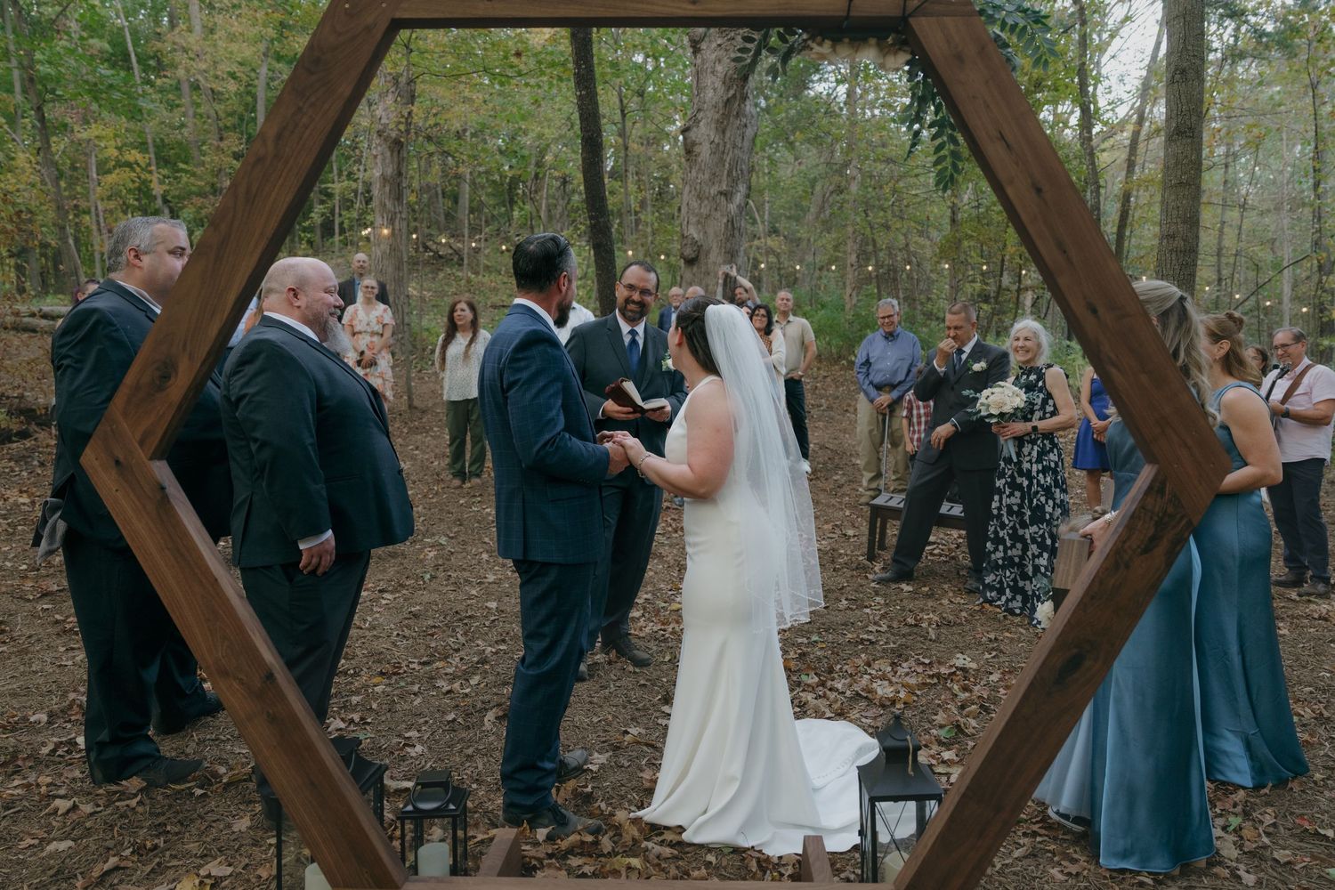 Outdoor woodland wedding ceremony under a hexagonal wooden arch surrounded by fall trees and guests in Shakopee, Minnesota.