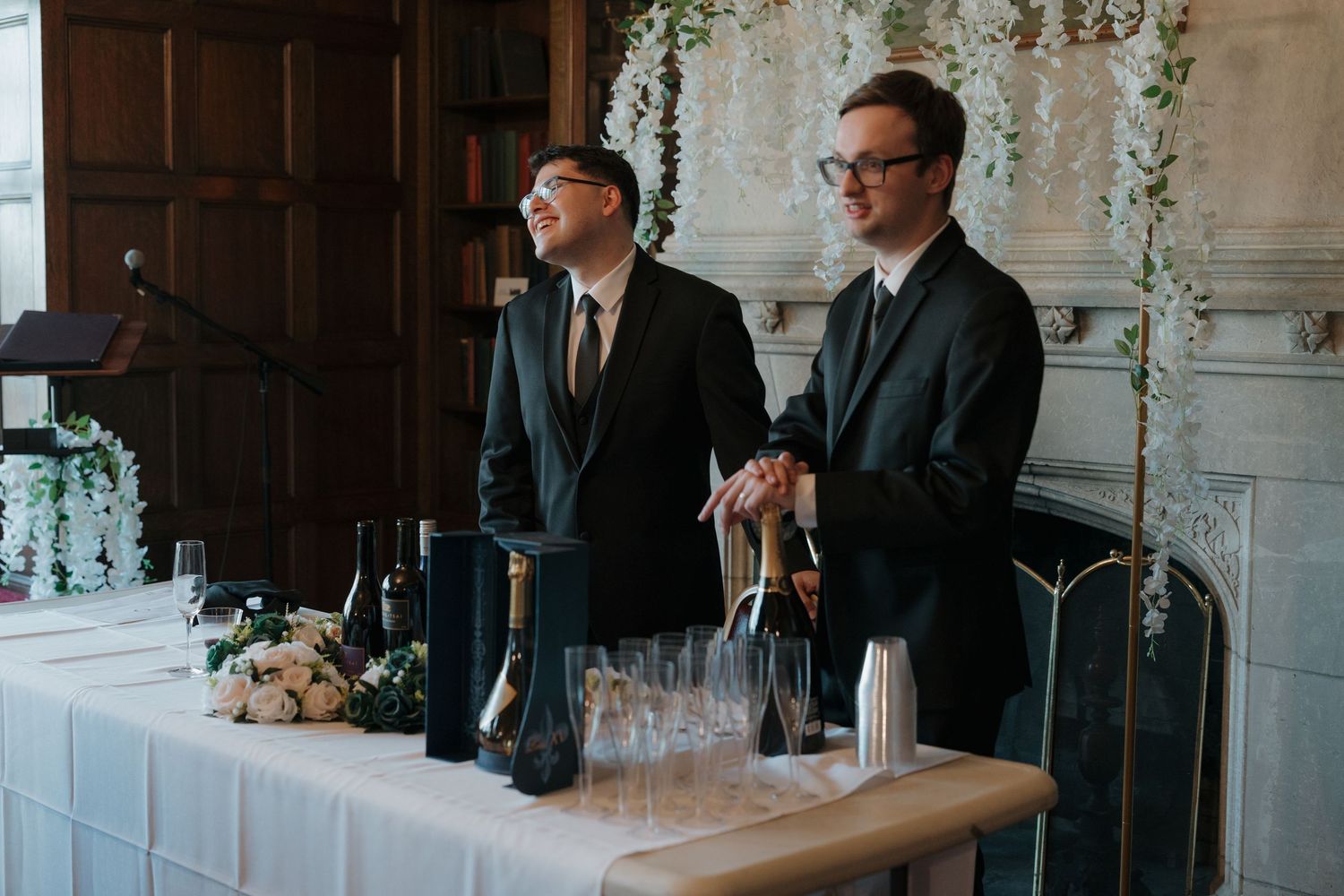 Two grooms in black suits stand at a reception table with champagne glasses and floral decor in Rochester, Minnesota at Plummer House.