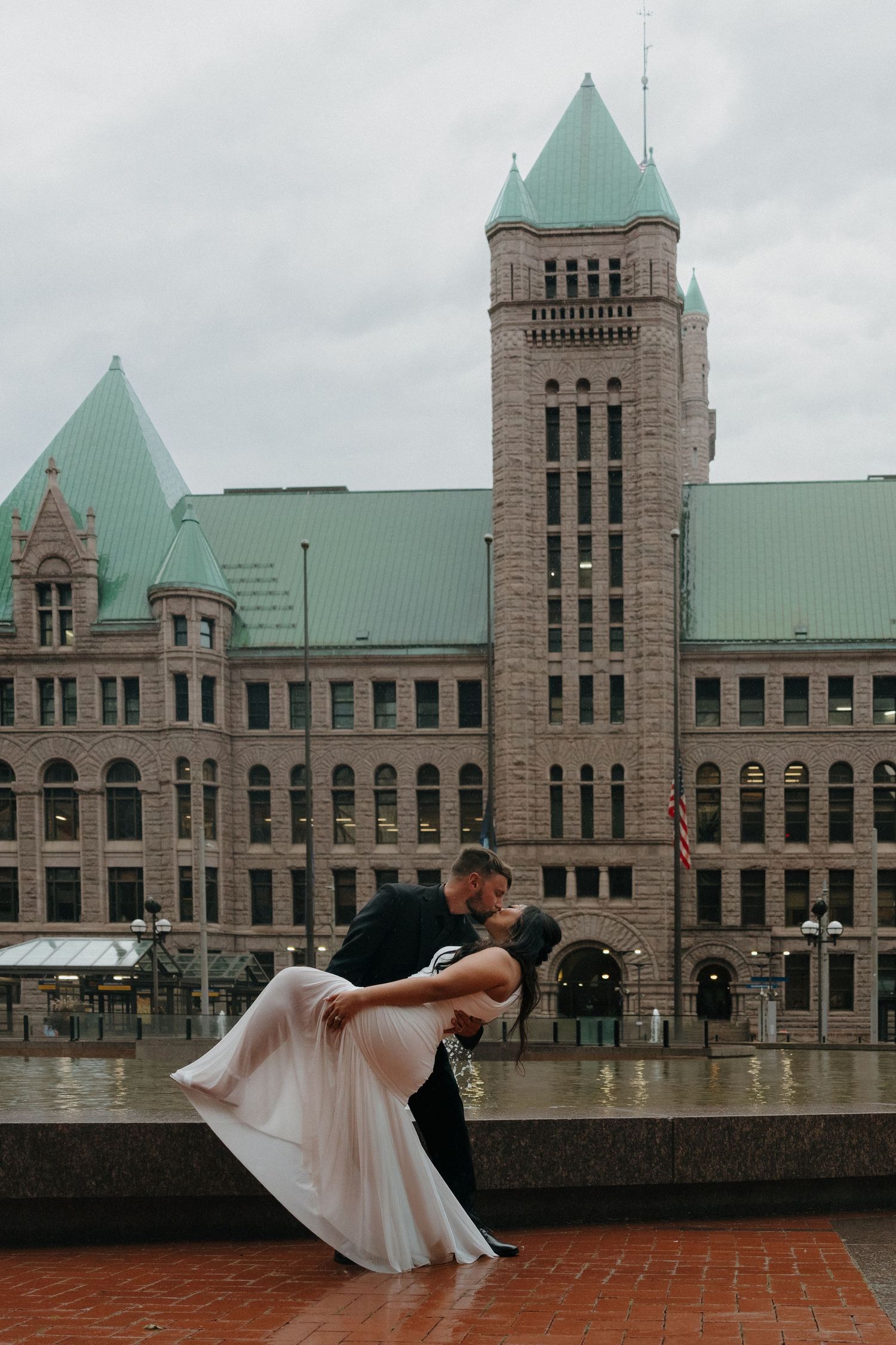 Groom dips bride dramatically in front of grand historic stone building on cloudy day outside Minneapolis City Hall.
