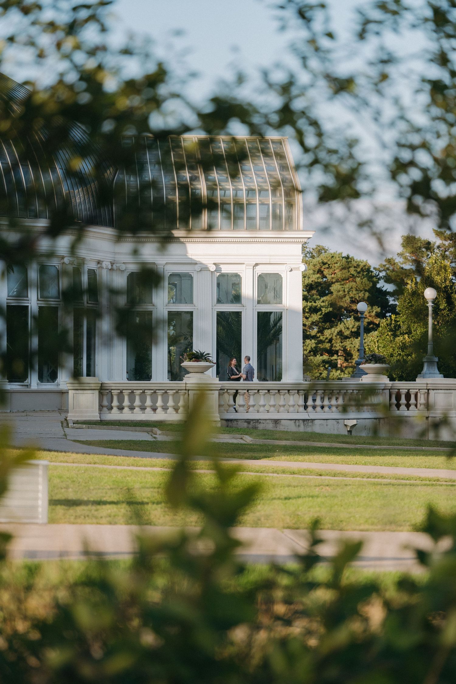 Couple sits on terrace of elegant white greenhouse conservatory of Como Conservatory in Saint Paul surrounded by lush greenery.