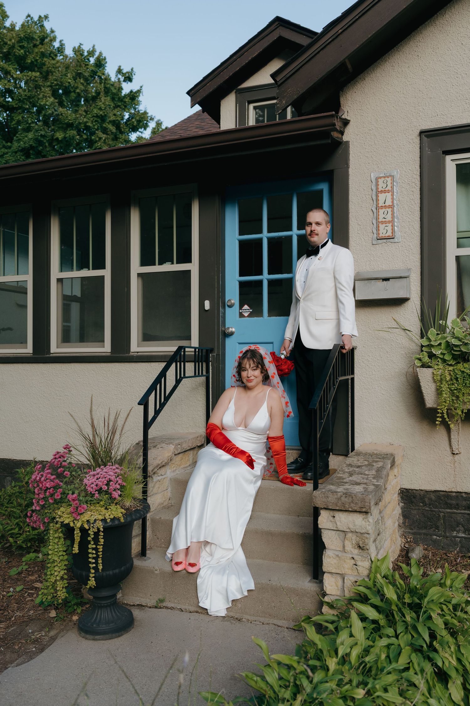 Bride in white gown with red gloves and groom in white suit pose on stone steps outside a house with blue door.