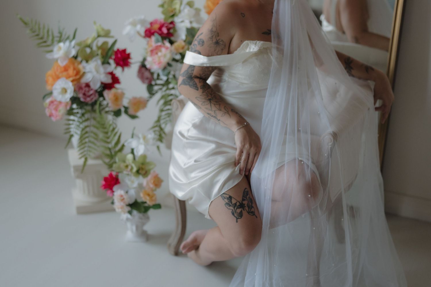 A tattooed bride in a white satin dress and veil kneels beside a vibrant floral wreath arrangement in a studio setting in Minneapolis, MN.