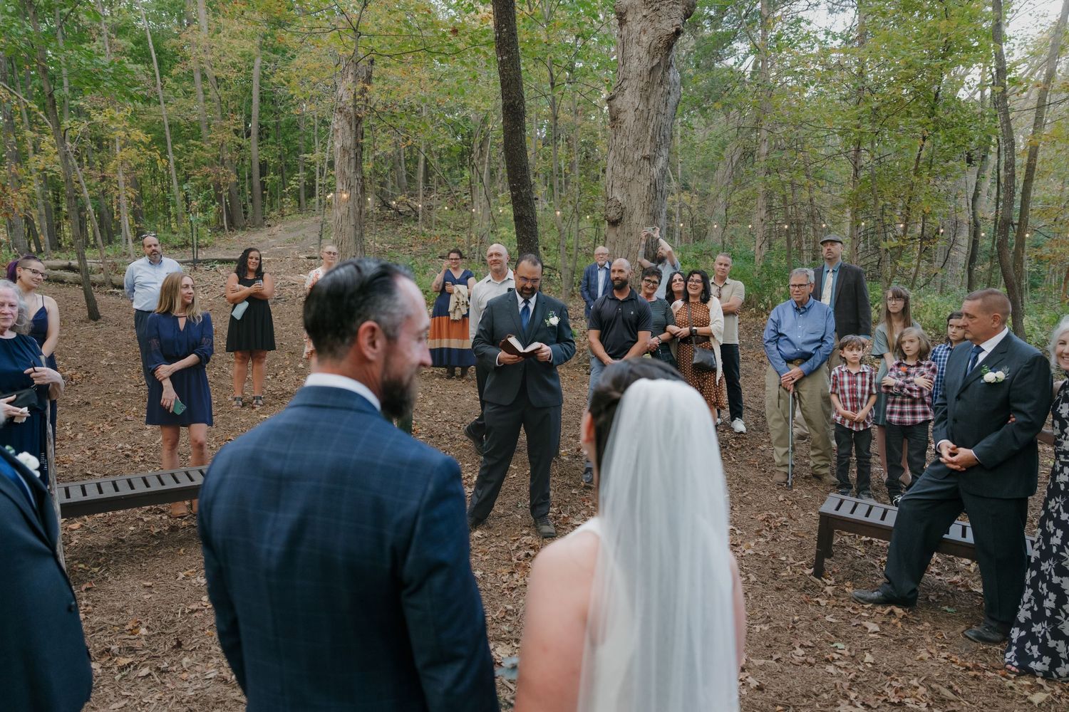 Outdoor woodland wedding ceremony with bride, groom, officiant, and guests among autumn trees in Southern Minnesota.