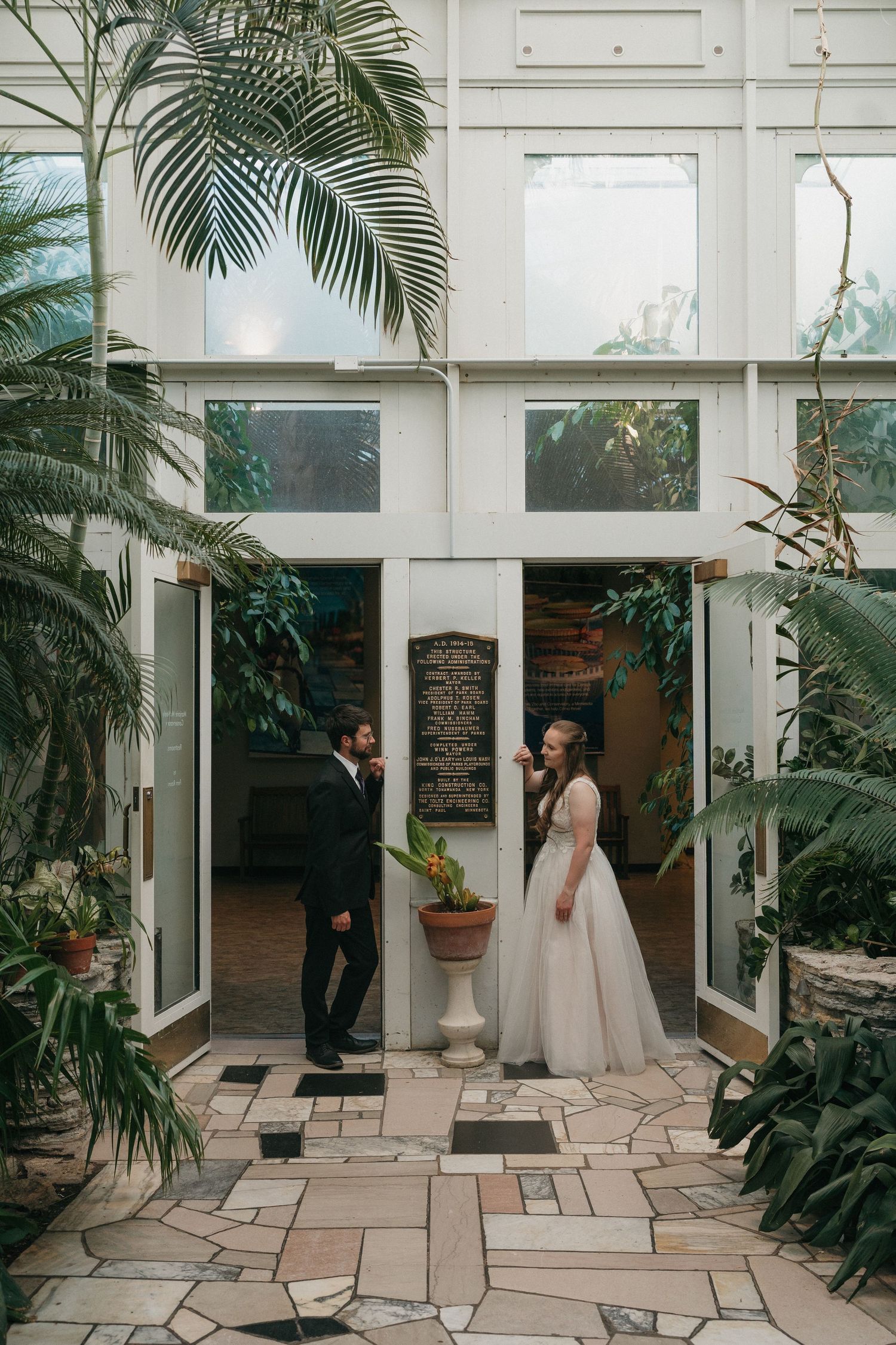 Bride and groom share a romantic moment at a lush greenhouse entrance, surrounded by tropical plants on their wedding day.