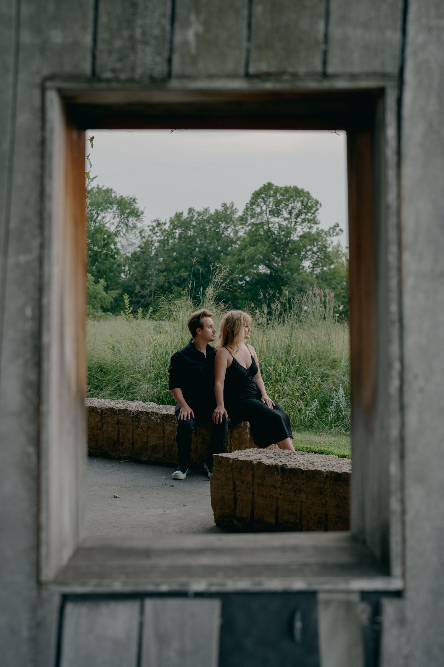 Couple sitting close together framed by a rustic wooden window opening with green meadow behind them in Hastings, Minnesota.