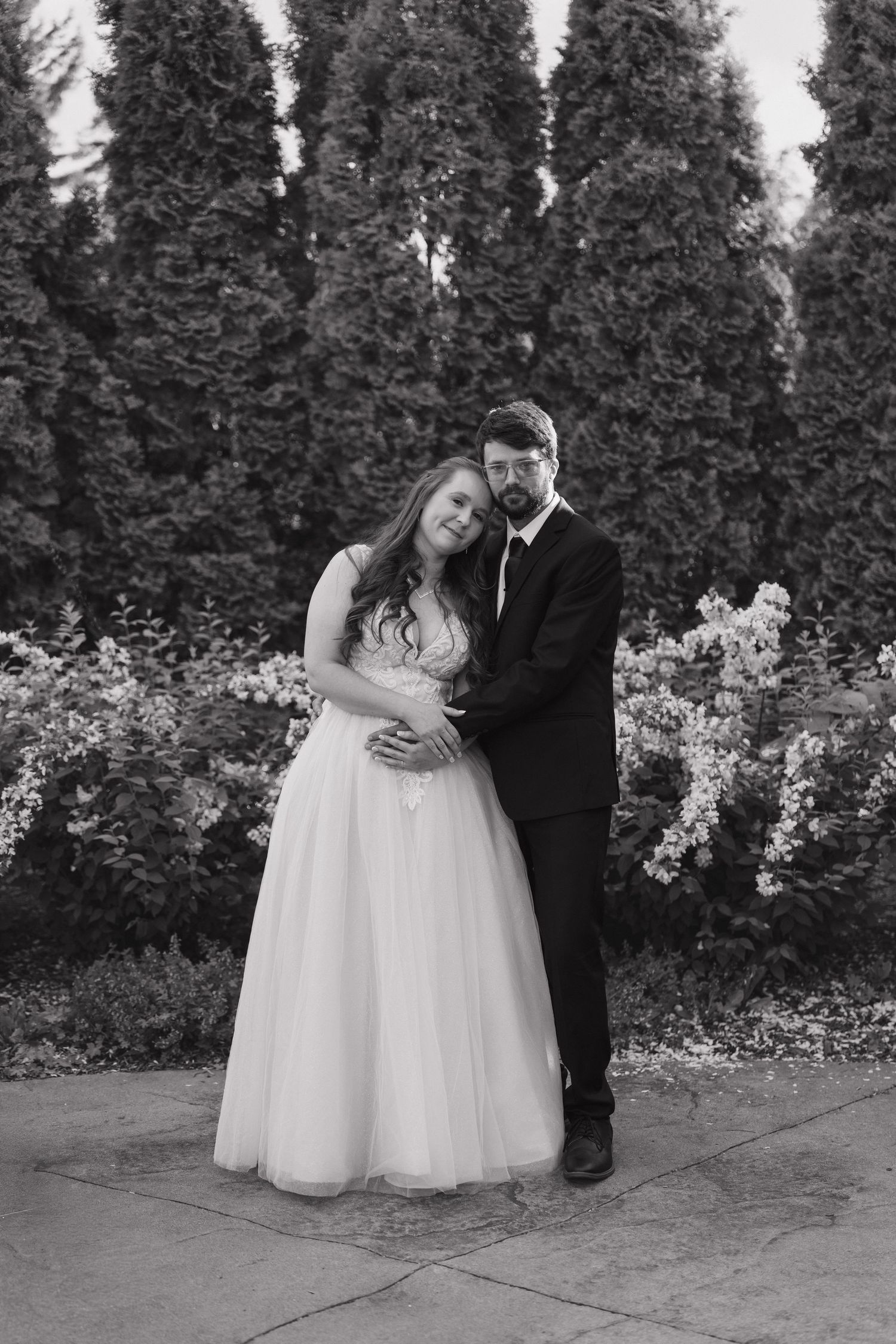 Black and white photo of a bride and groom embracing in a garden, surrounded by tall evergreen trees.