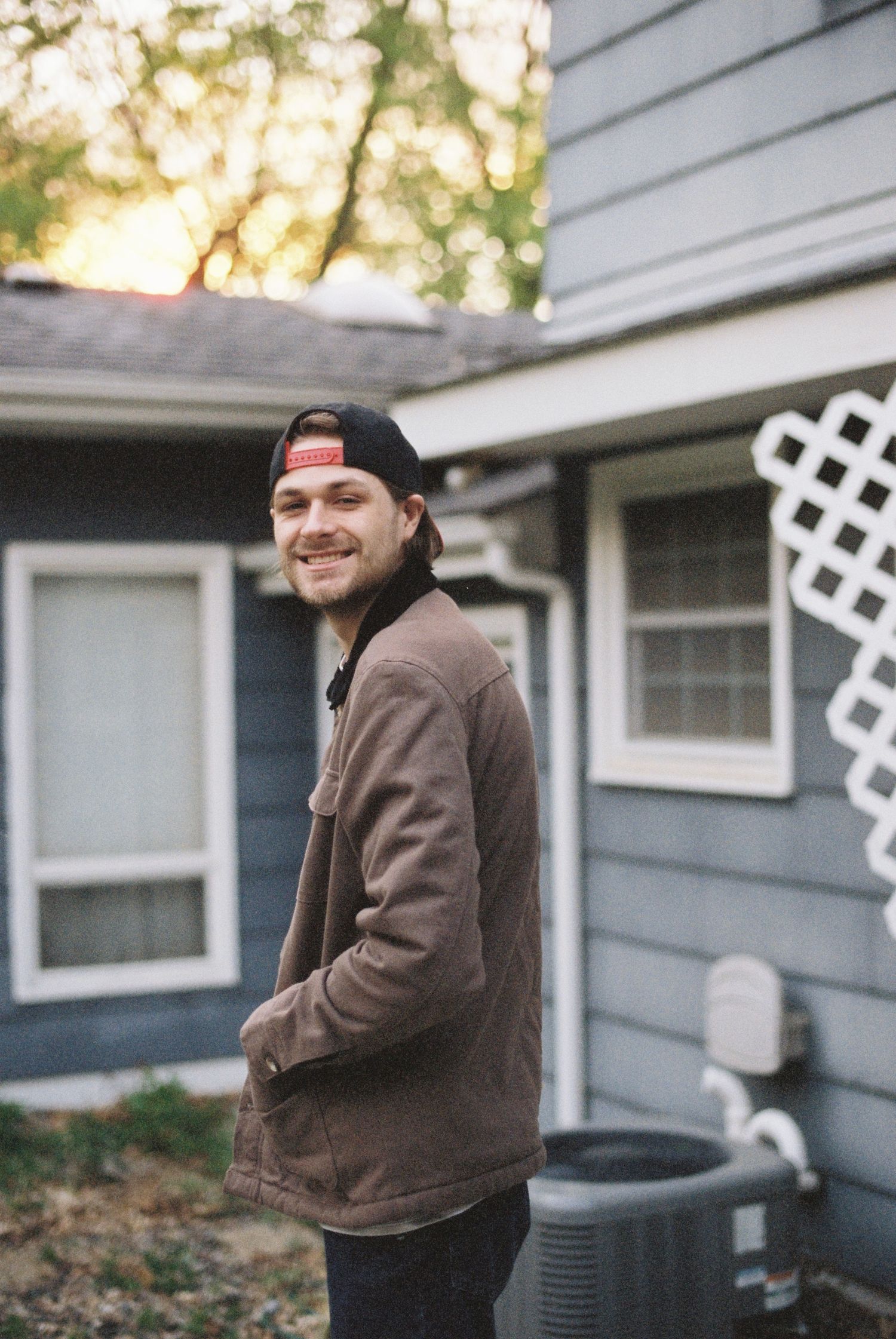 Smiling young man in a brown jacket and baseball cap standing outside a blue house at dusk in Minnesota.