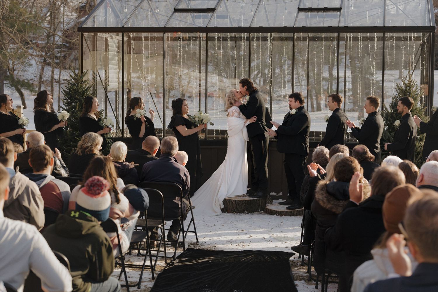 Couple shares first kiss at outdoor winter wedding ceremony with guests seated in rows before a greenhouse backdrop in Minnesota.