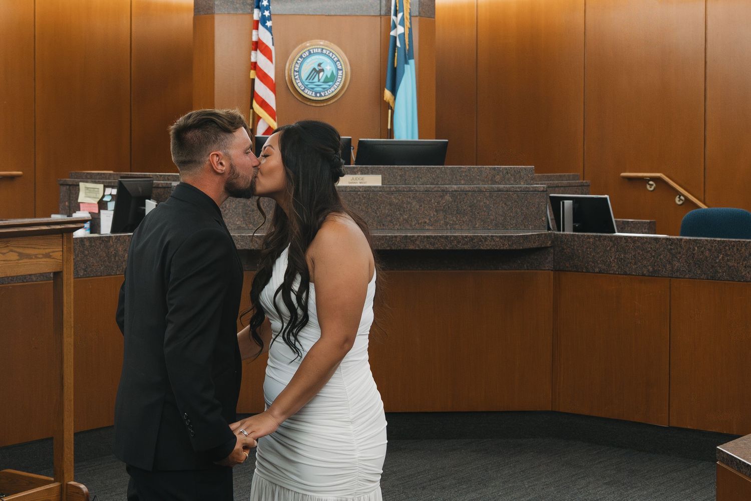 Couple sharing a kiss at courthouse wedding ceremony in formal attire, with American flag and official seal in background.