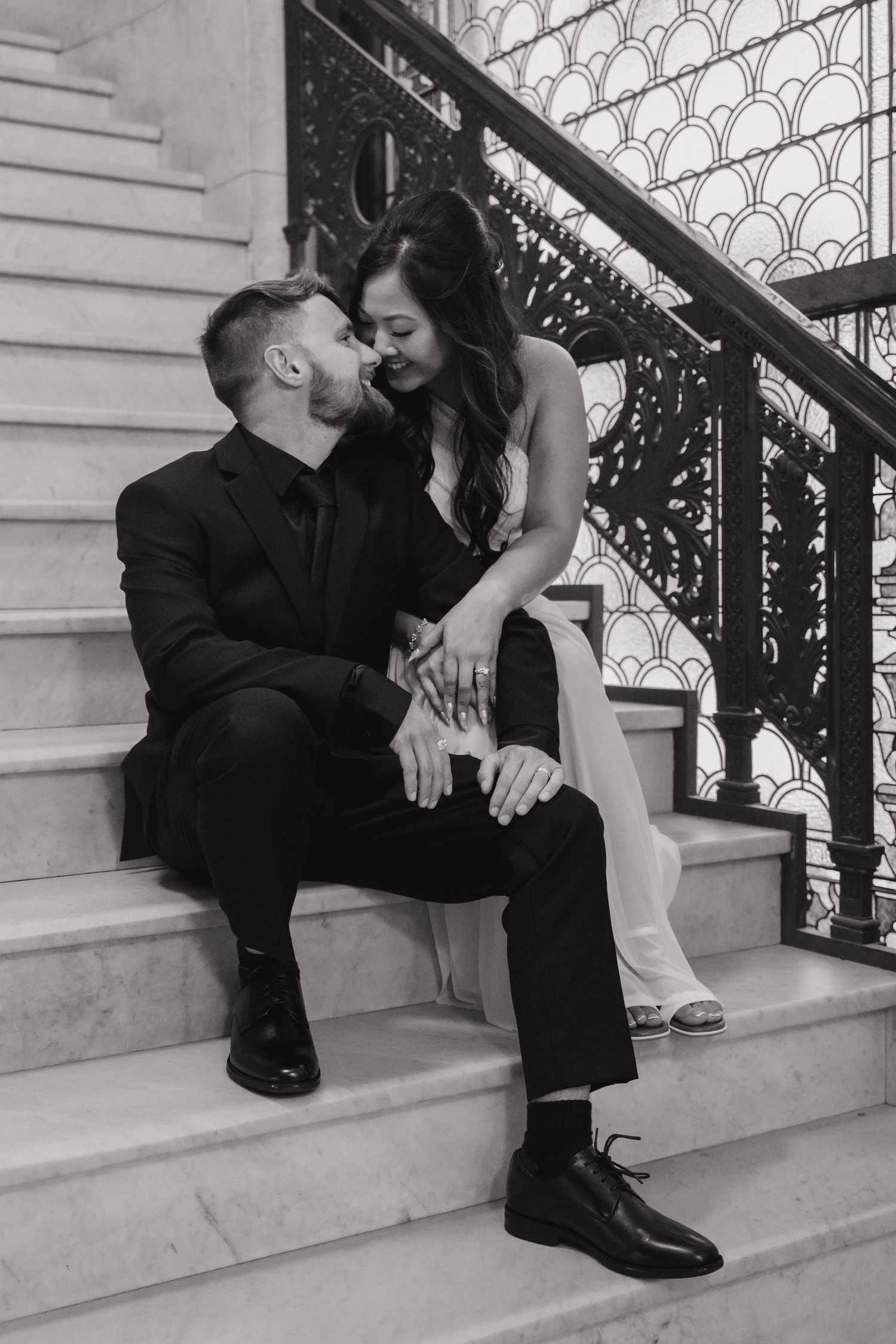 Black and white photo of a couple sitting on marble stairs, sharing a romantic moment in formal attire.