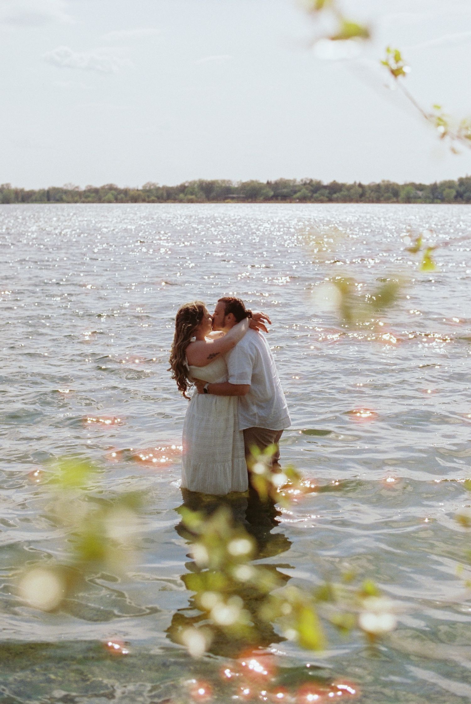 A couple shares a romantic kiss while standing in a shimmering lake, framed by soft green leaves in golden light.