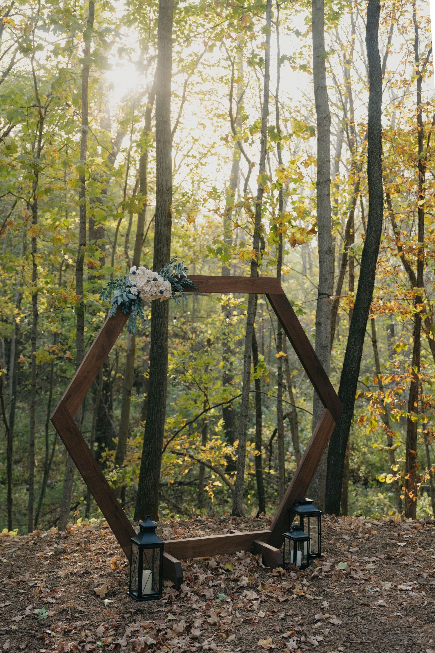 Rustic hexagonal wooden wedding arch decorated with flowers and lanterns in a sunlit autumn forest in Southern Minnesota.