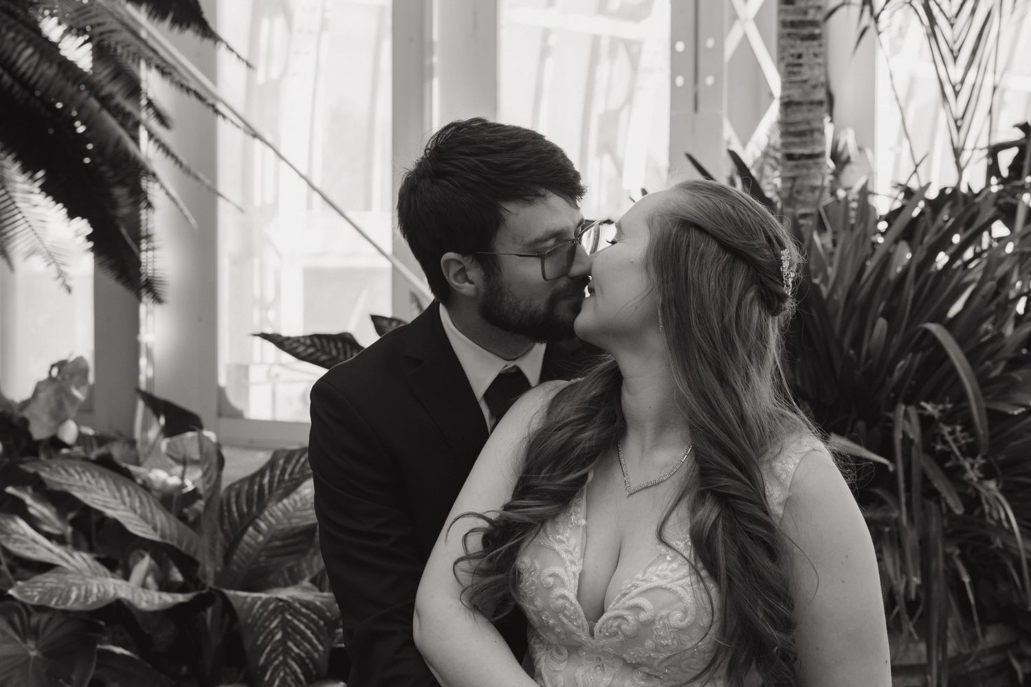 Black and white photo of a couple sharing a romantic kiss at their wedding surrounded by lush tropical plants.