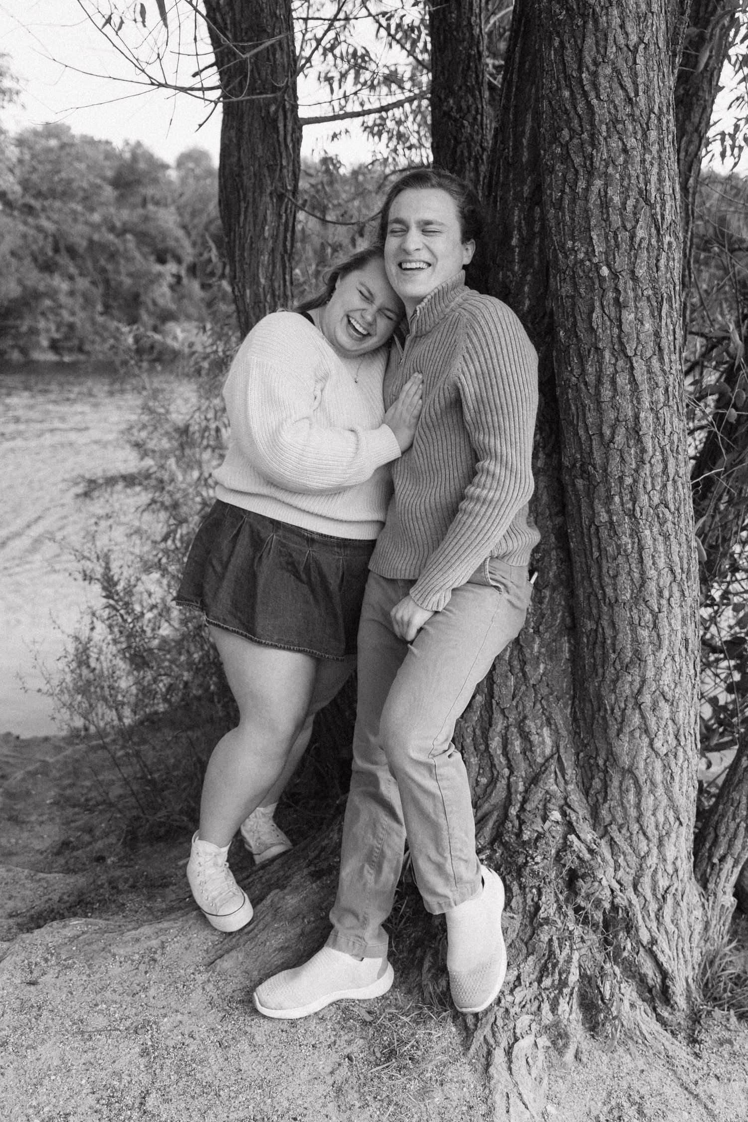 Black and white photo of laughing couple leaning against trees near a river in a natural setting at a local park.